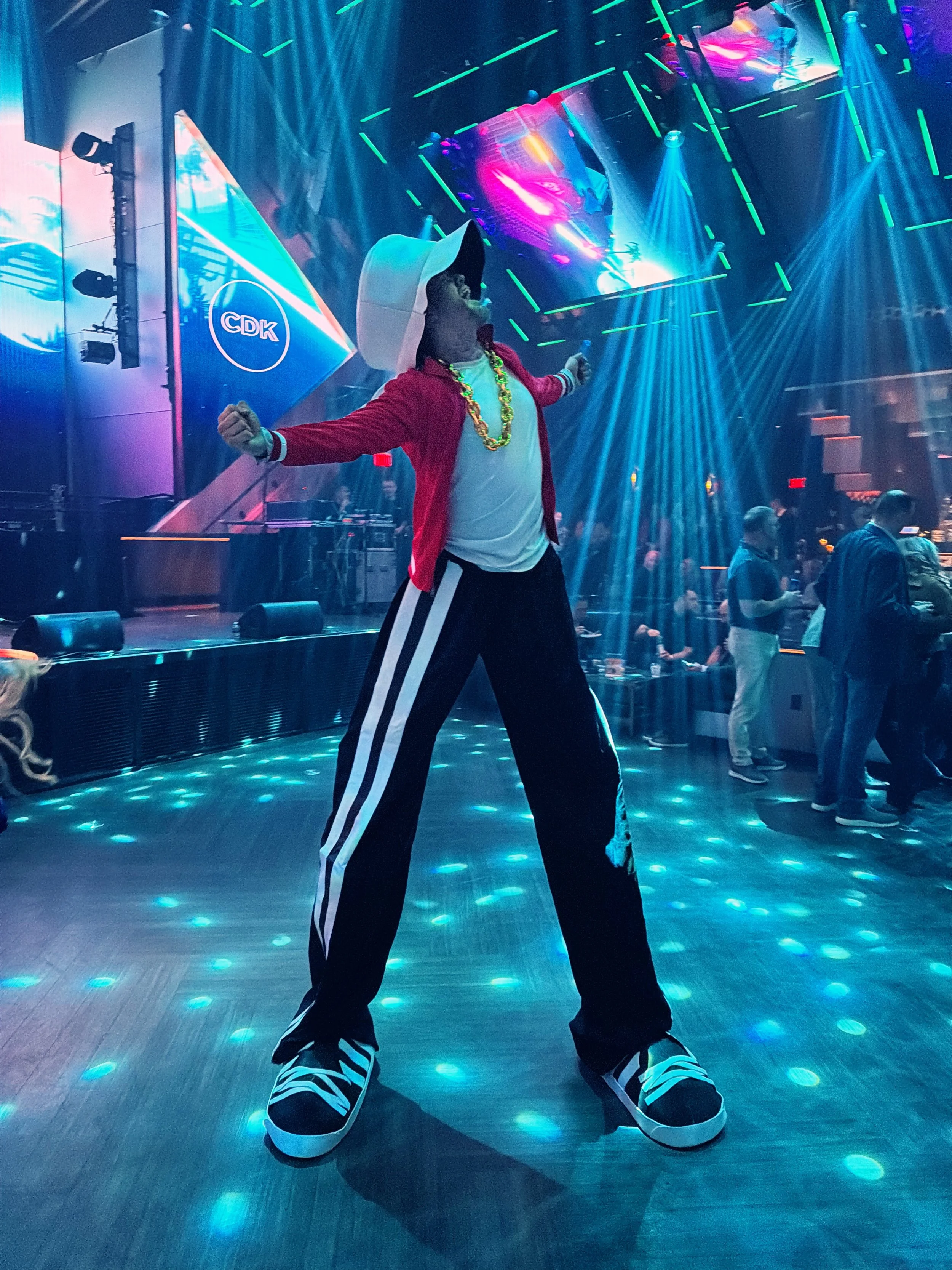 A person wearing a large white hat, red jacket, white T-shirt, black and white striped pants, and oversized shoes is dancing in a nightclub with colorful lights and a DJ booth in the background.