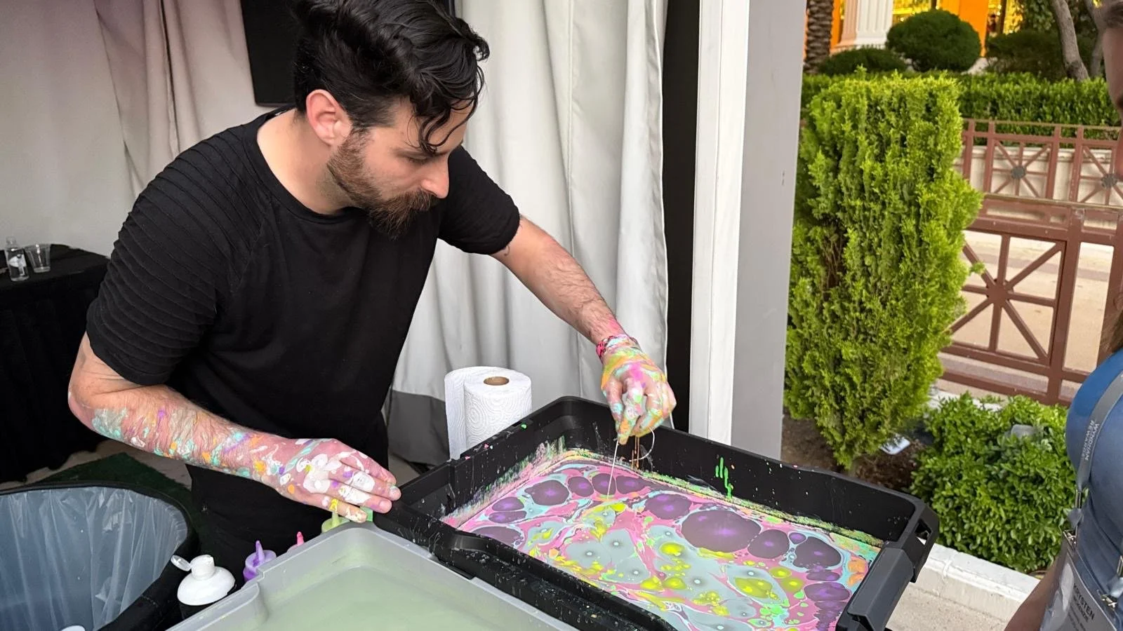 A man with dark hair and a beard creating colorful soap bubbles in a tray, wearing a black shirt and disposable gloves, standing outside next to green bushes and a decorative fence.