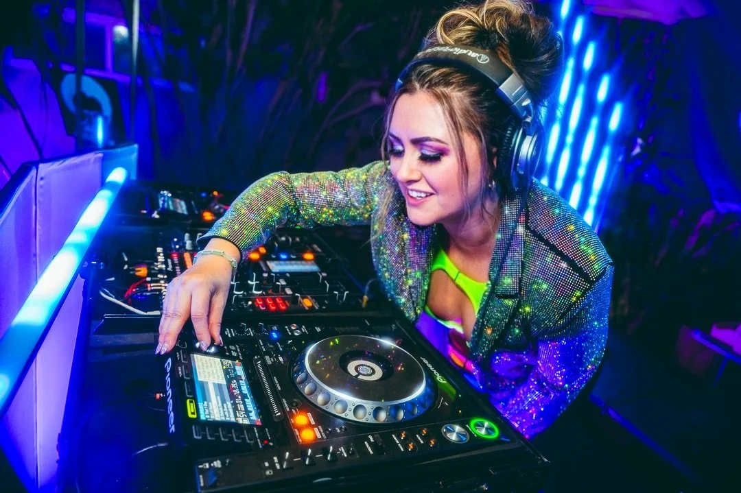 A female DJ wearing headphones and a shimmering jacket, smiling while mixing music on a DJ controller in a nightclub with blue and purple lighting.