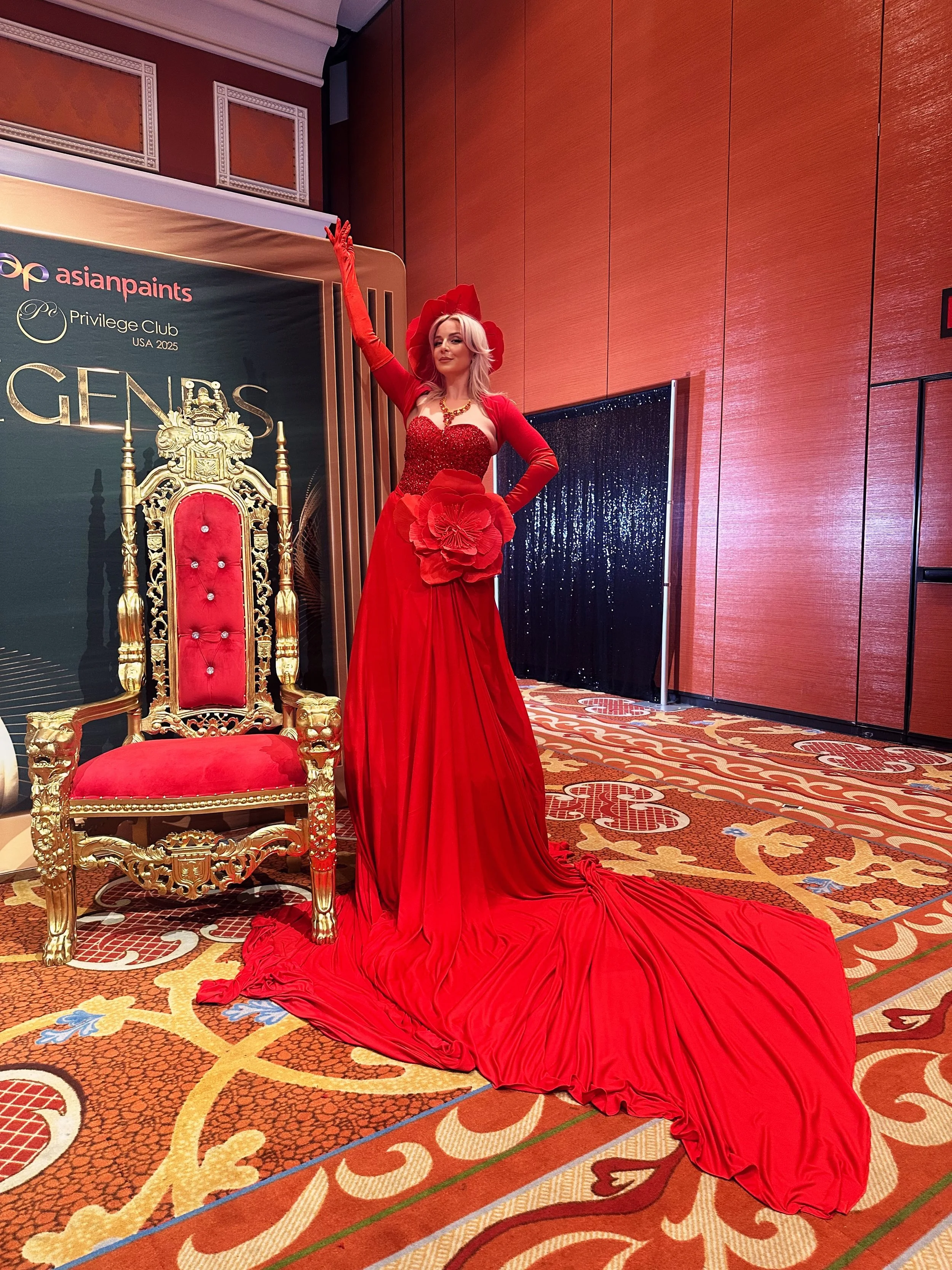 A woman dressed in an elaborate red gown with a large fabric flower on the waist, standing beside a gilded throne. She is posing with one arm raised and wearing matching red gloves and a hat. The backdrop features a black and gold banner with the log