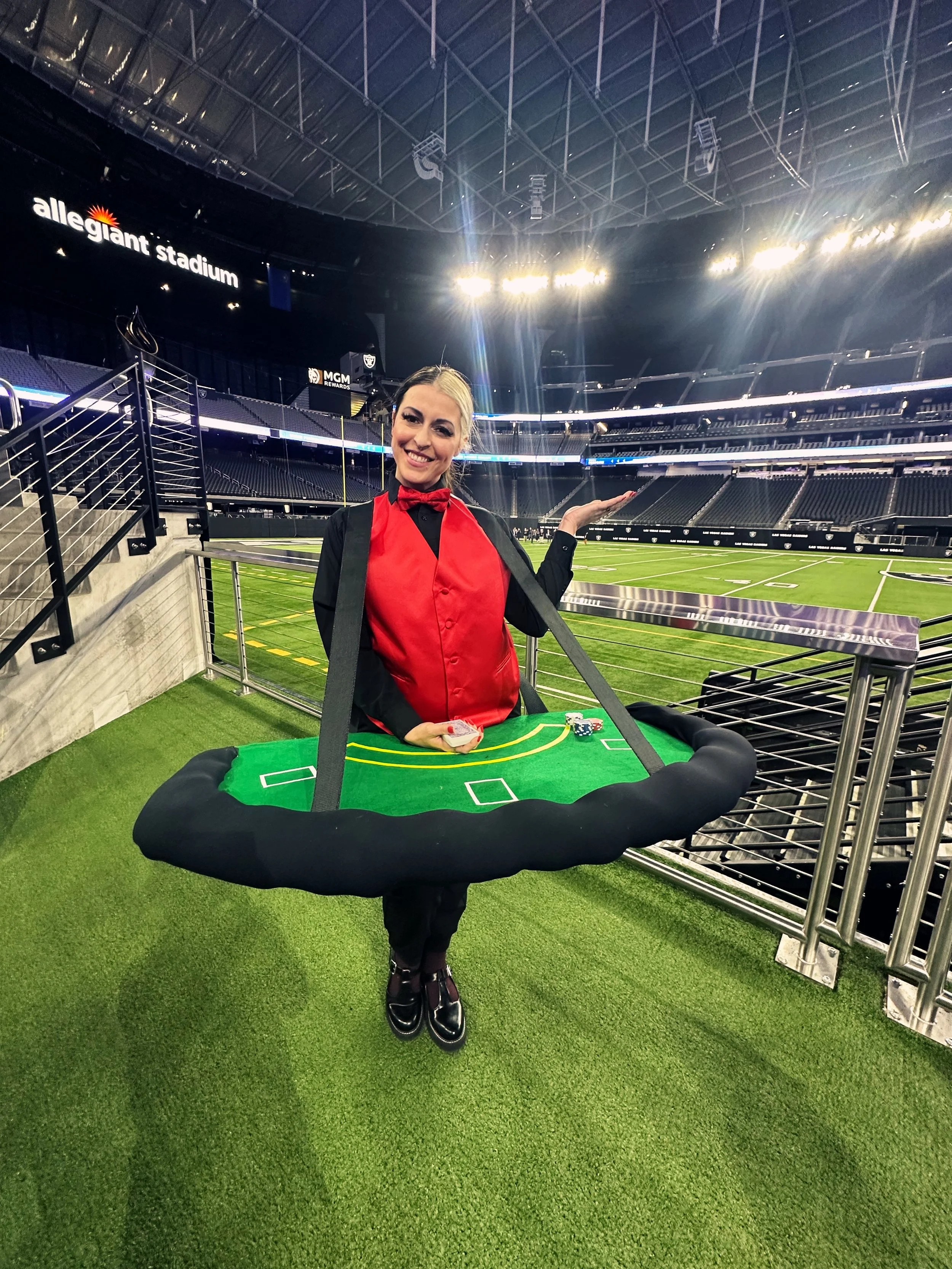 A woman dressed as a hostess at Allegiant Stadium, standing on a green artificial turf with a miniature football field around her waist, smiling and posing inside the stadium.