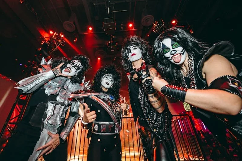 Four people dressed as members of KISS in stage makeup and costumes, standing on a concert stage under red lighting.