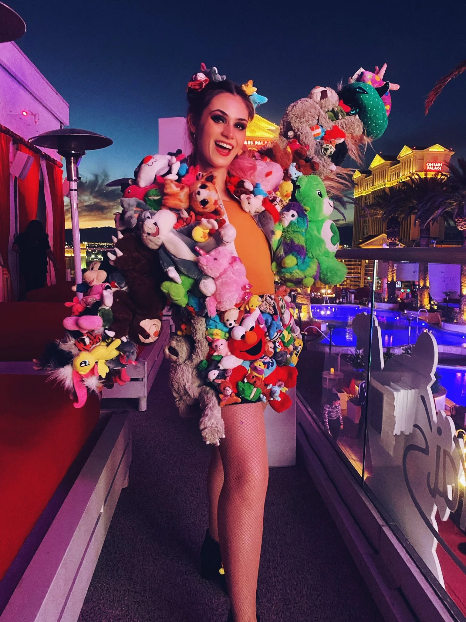 Woman smiling, wearing a jacket decorated with colorful plush stuffed animals, in an outdoor setting at dusk with city lights, a swimming pool, and illuminated buildings in the background.