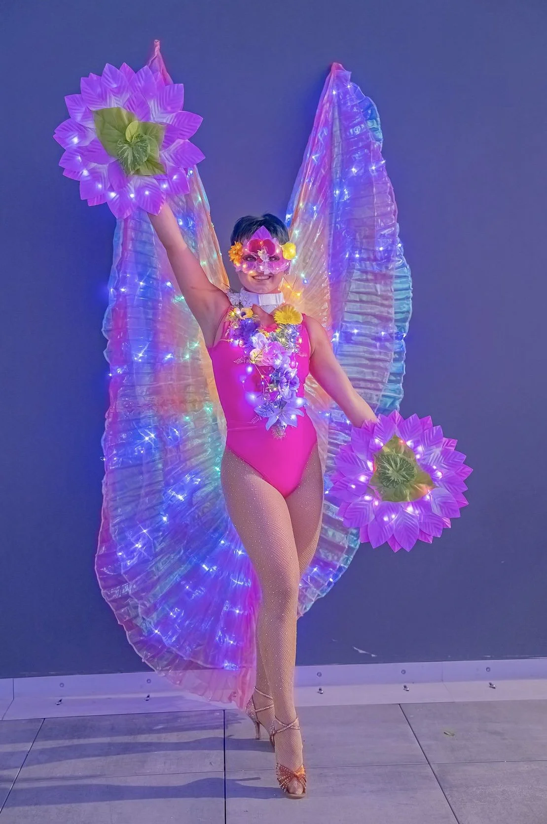 Performer in vibrant costume with LED lights, colorful flower accessories, and large spiked wings, holding oversized purple flowers against a dark background.