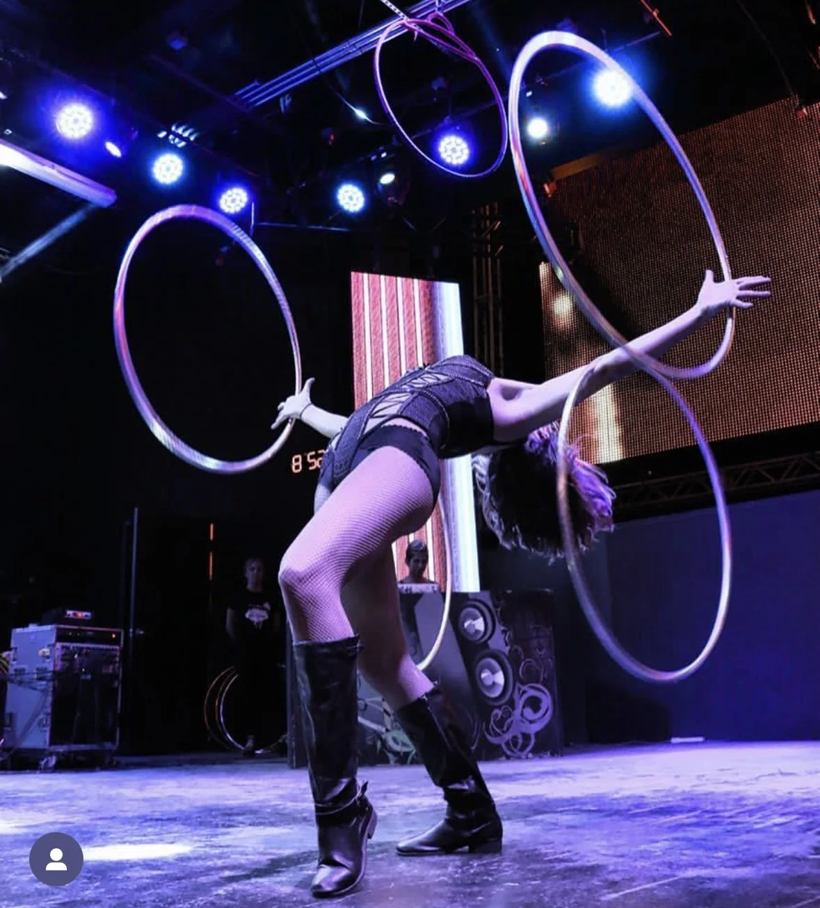 A female performer with short hair in black outfit and high boots performing a contortion act with colorful hoops on stage under purple and blue lights.