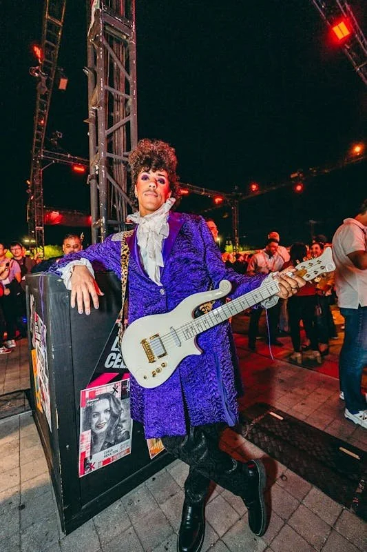 Person dressed in a vibrant purple costume holding a white electric guitar at an outdoor event during nighttime.