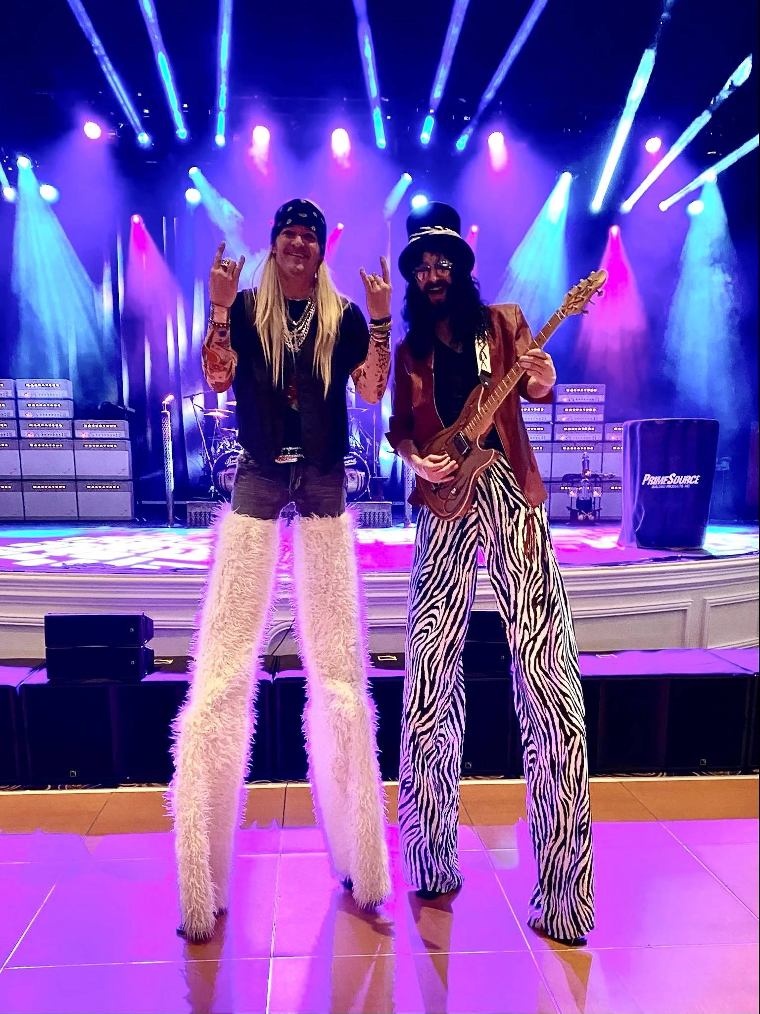 Two musicians on stage with vibrant purple and blue lighting, one with long blonde hair, wearing fluffy white pants and a bandana, and the other with long dark hair, beard, glasses, a hat, striped pants, holding a guitar.