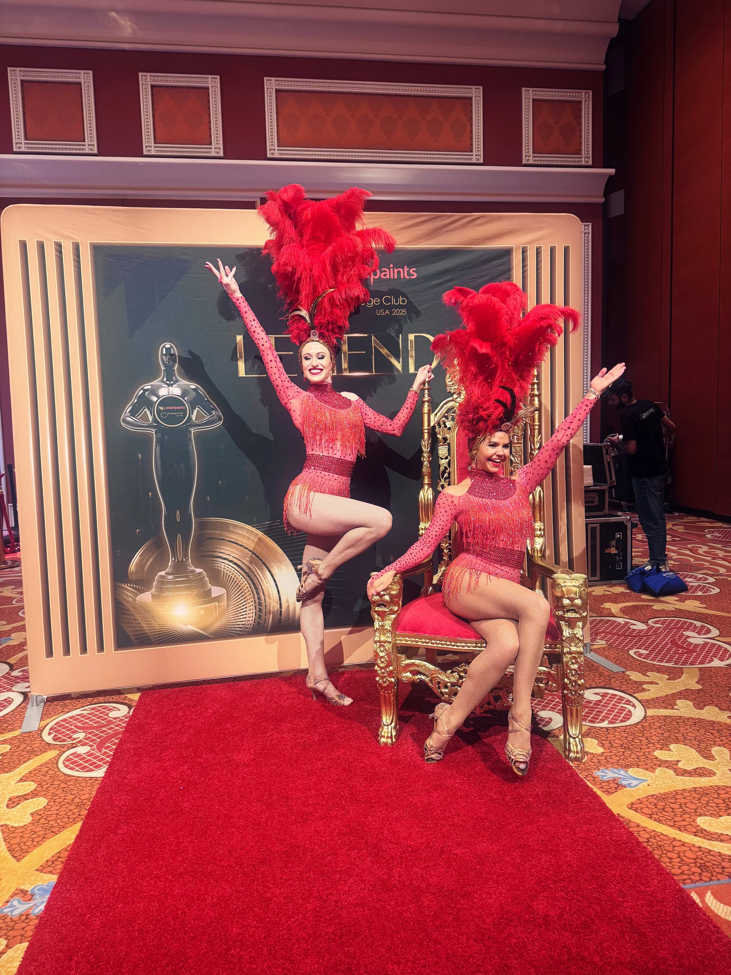 Two women dressed in red showgirl costumes with large feather headdresses, posing on a red carpet in front of a backdrop with an award statue and text.