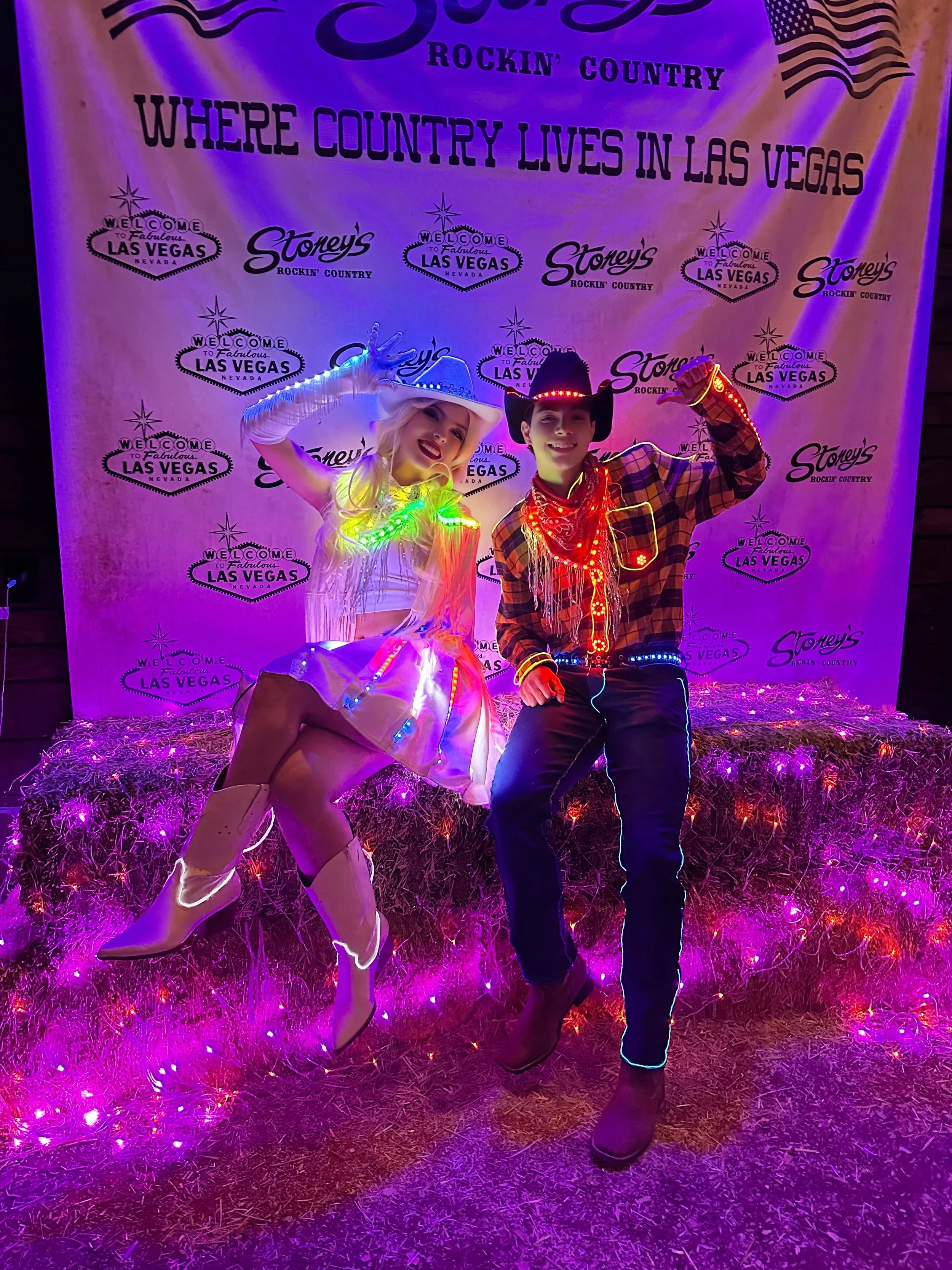 Two people dressed in illuminated cowboy and cowgirl costumes, sitting on hay bales with purple and pink lights, against a backdrop that reads 'Where country lives in Las Vegas' and 'Rockin' country'.