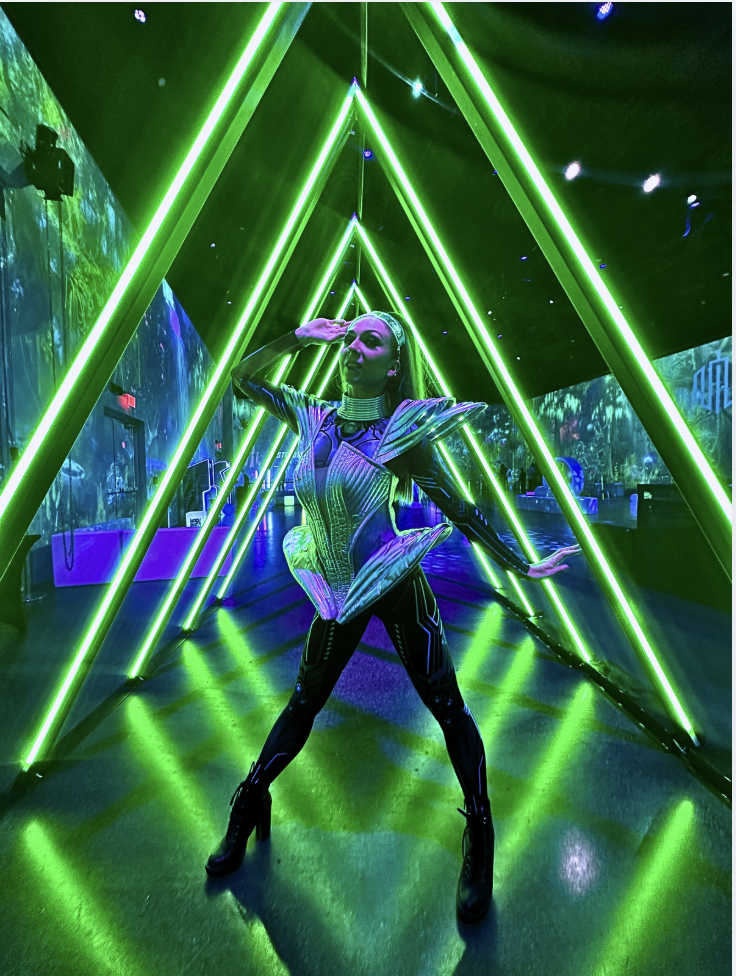 A woman in a futuristic, metallic outfit posing inside a tunnel of green laser lights with neon reflections and digital screens in the background.
