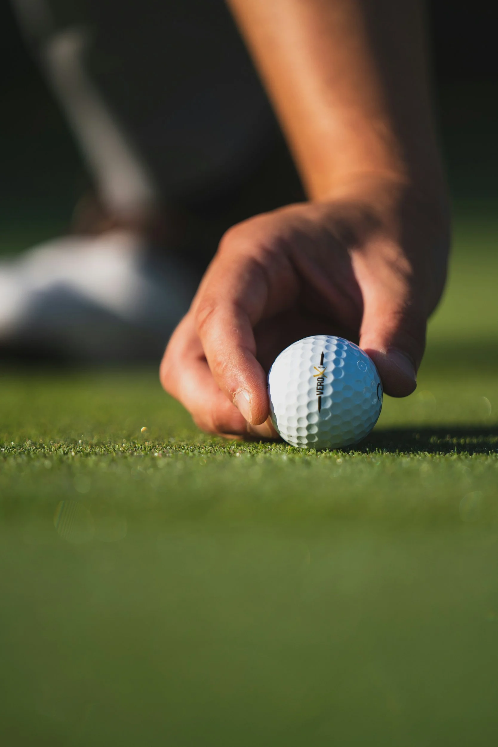 Close-up of a person's hand placing a golf ball on the grass, with a focus on the golf ball and hand.