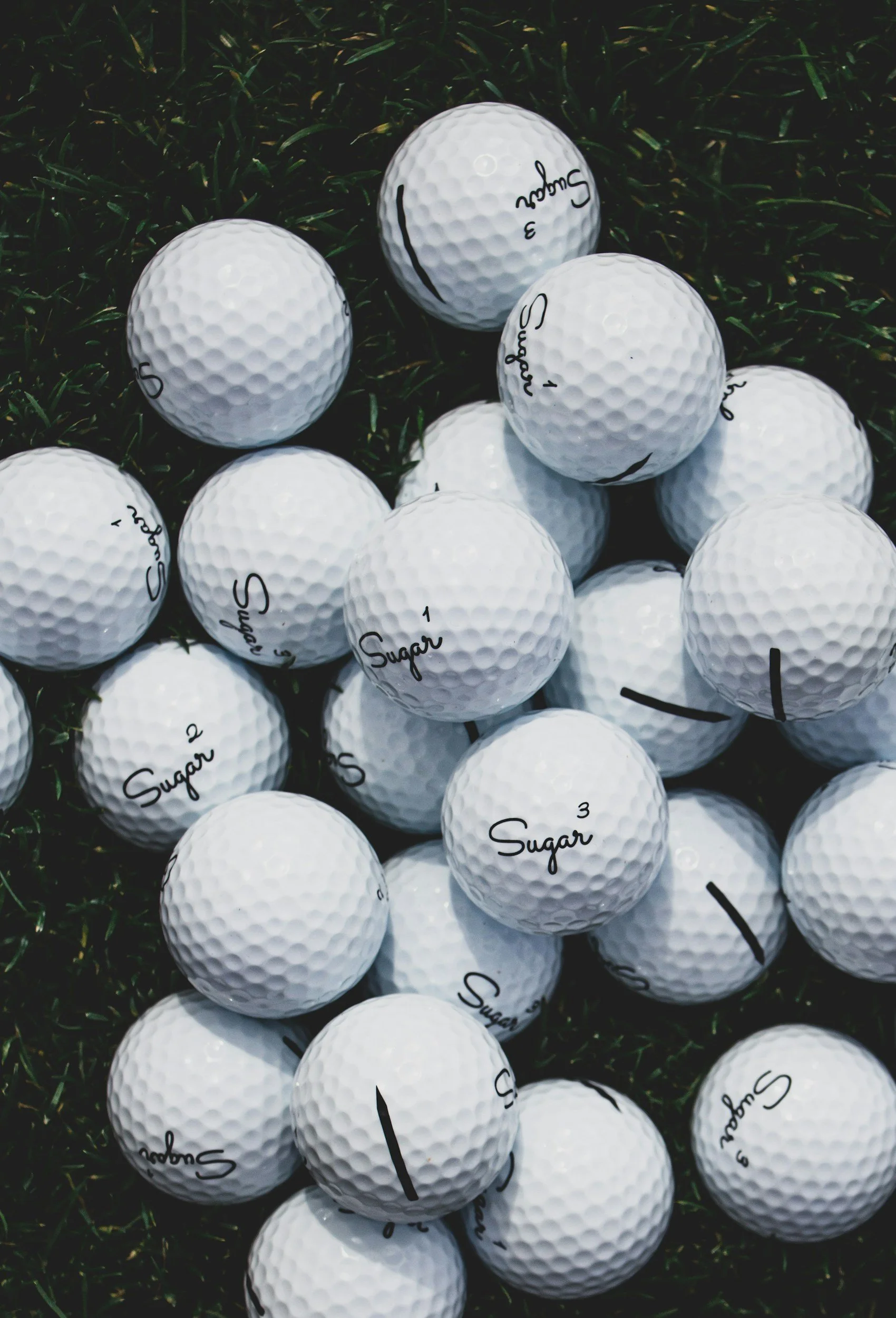 A pile of white golf balls with the word 'Sugar' and numbers written on them, resting on green grass.