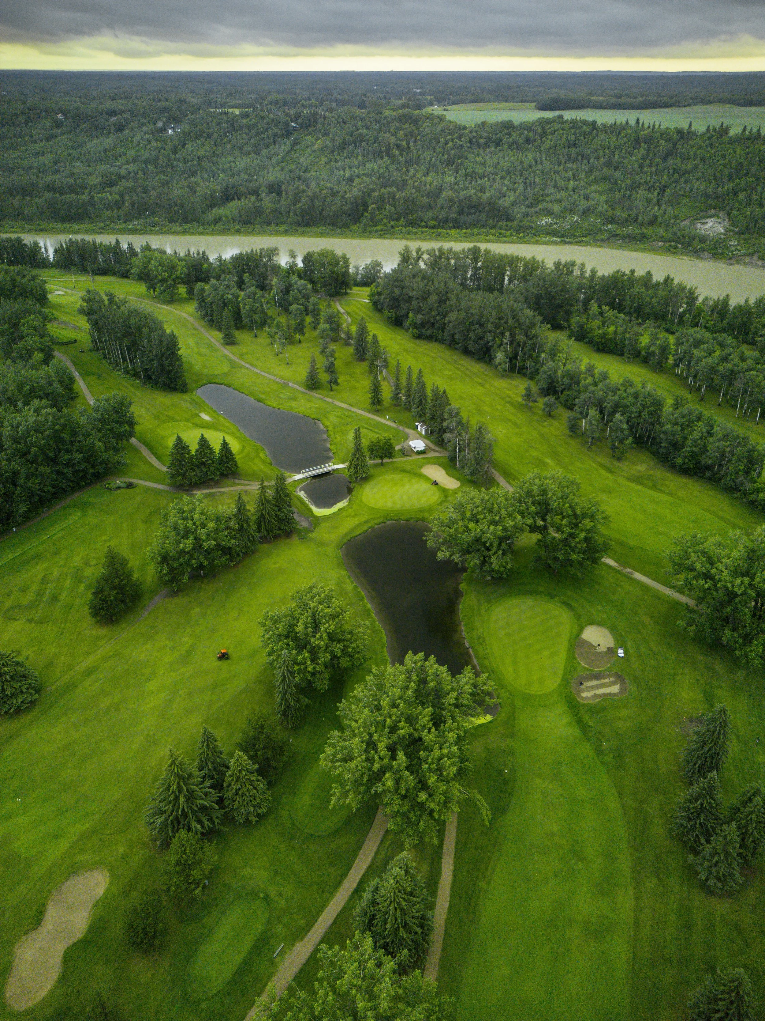Aerial view of a lush green golf course with sand traps, water hazards, and a small bridge over a pond, surrounded by trees and dense forest, with cloudy skies overhead.