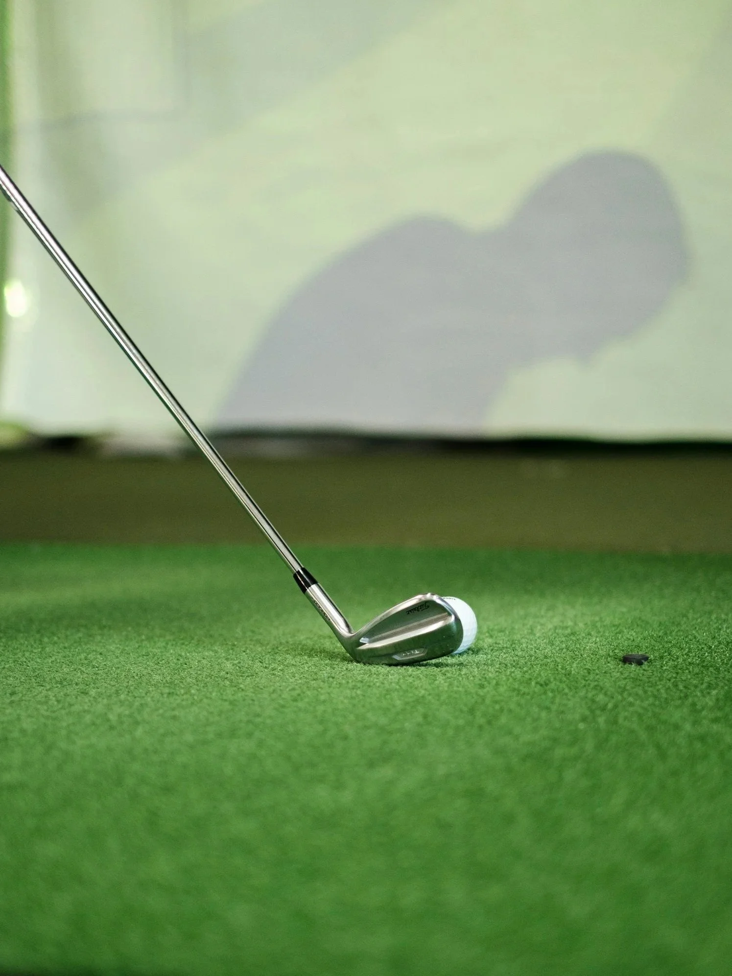 Golf club positioned behind a golf ball on a green putting surface, with the club's shadow cast on the background surface.