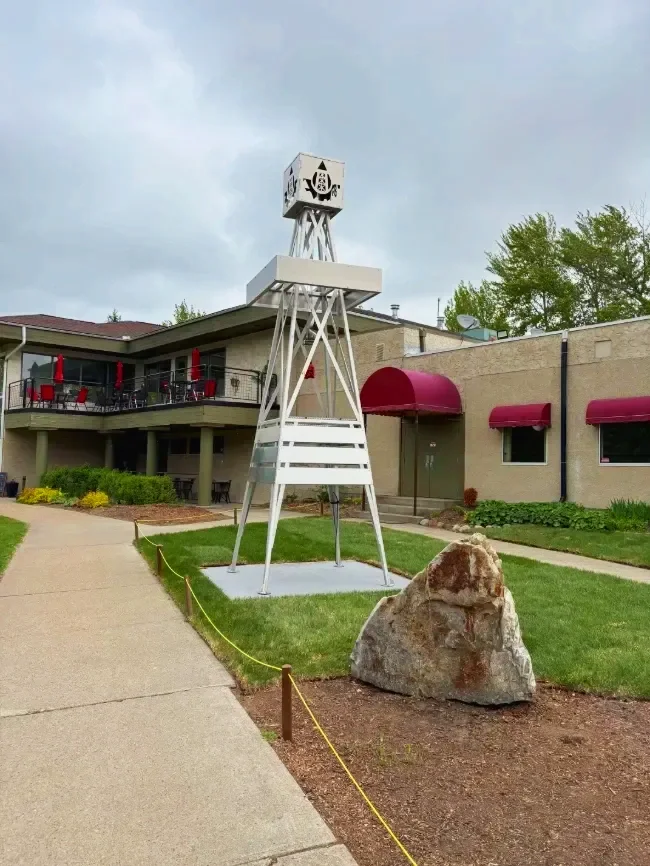 A tall, white, tripod-style windmill sculpture with a small, enclosed cabin at the top featuring a nautical emblem, situated outdoors on a small patch of grass with a large rock nearby, in front of a building with a balcony and red awnings.