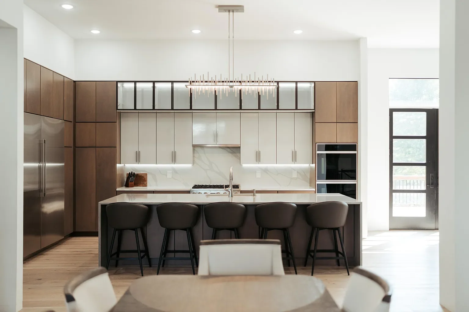 Modern kitchen with wooden and white cabinetry, marble backsplash, island with seating, stainless steel refrigerator, double ovens, and a chandelier above the island, with a door leading outside.