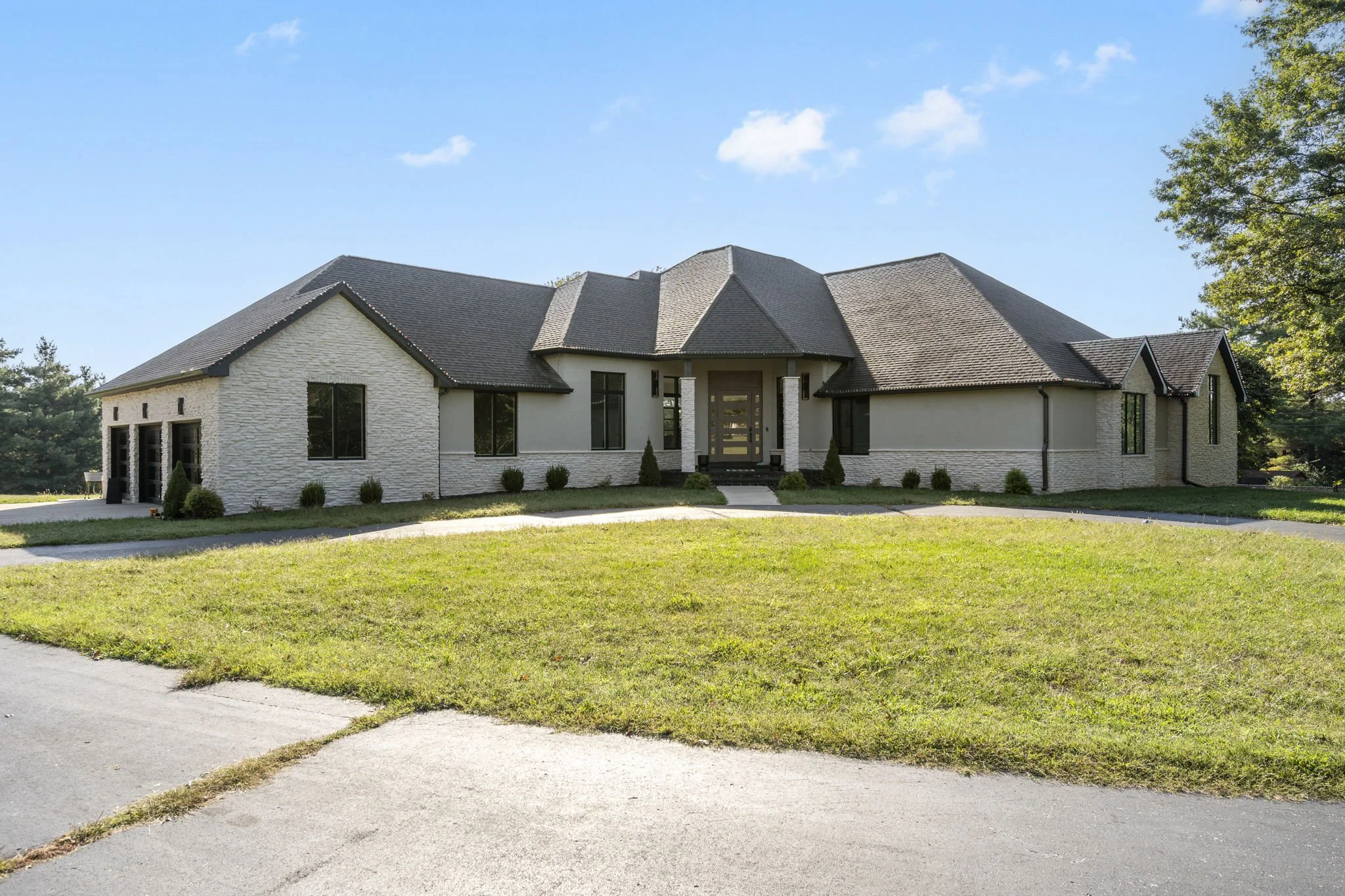 A modern, large single-family house with a sloped roof, white brick exterior, multiple windows, and a central front door with glass panels. The house is surrounded by a grassy lawn and a curved concrete driveway.