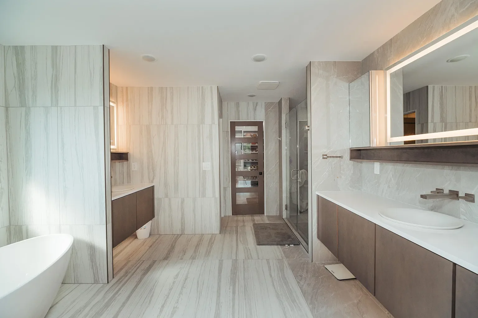 Modern bathroom with light-colored marble walls and floors, brown vanity with white countertop, large illuminated mirror, glass shower enclosure, and bathtub.