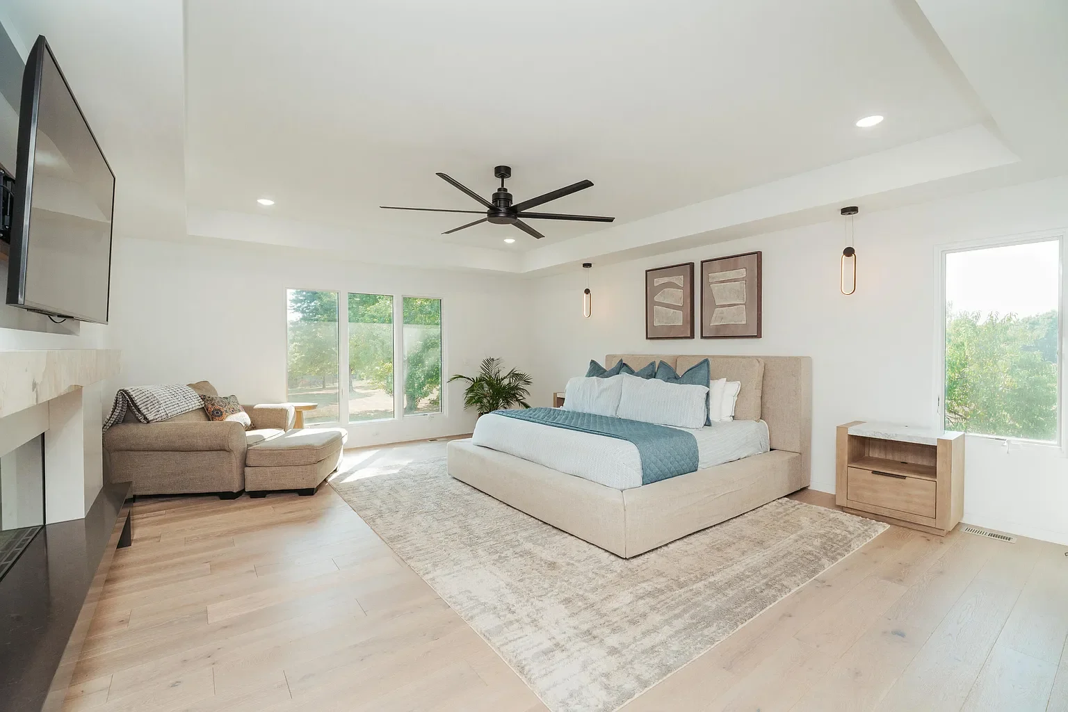 Modern bedroom with large bed, beige upholstered headboard, blue bedding, two framed abstract artworks, nightstand, ceiling fan, pendant lights, sitting area with beige chaise lounge, window views of greenery, light hardwood floors, and neutral decor.