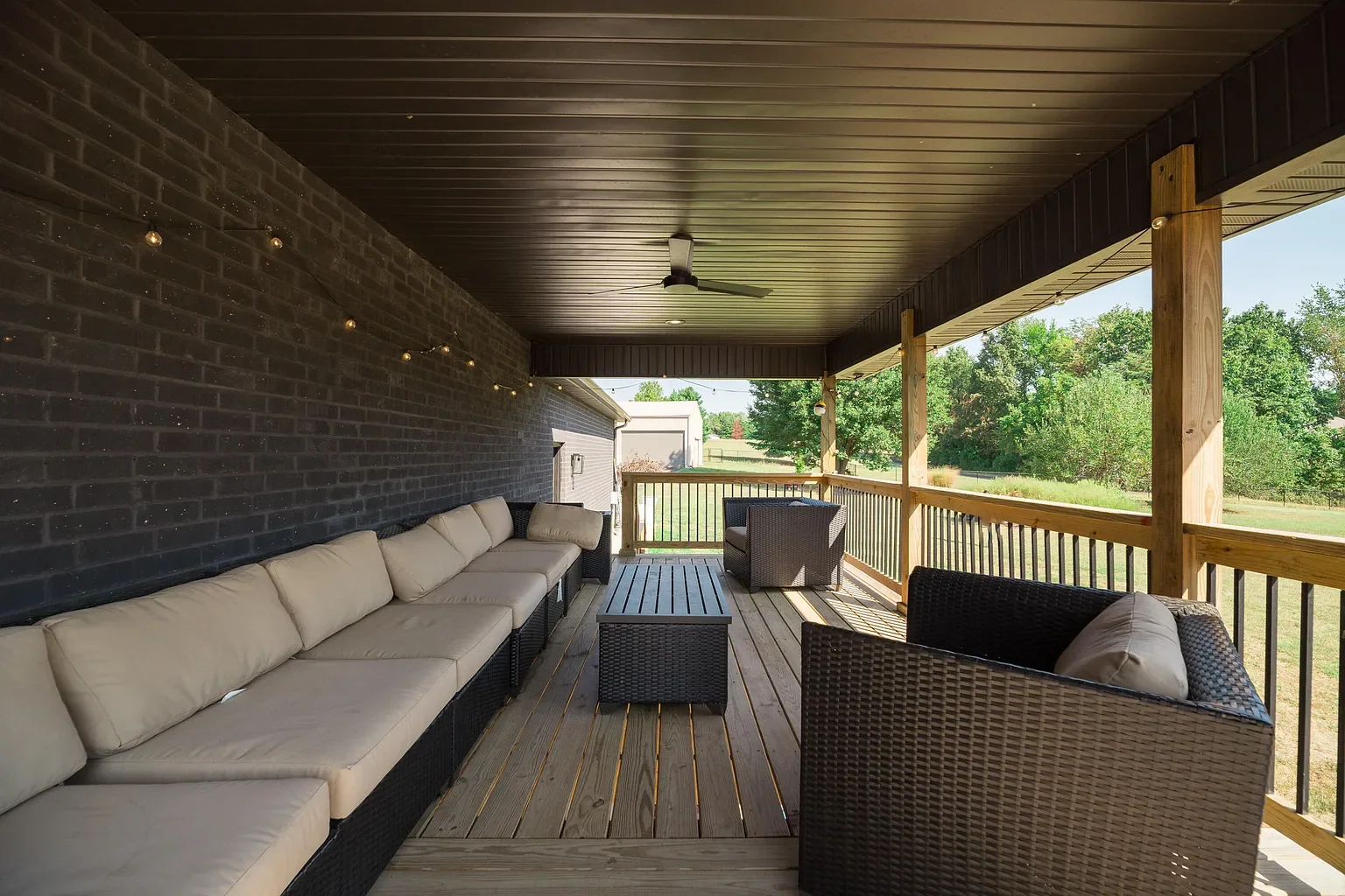 Covered outdoor porch with wicker furniture and string lights, overlooking a grassy yard with trees.