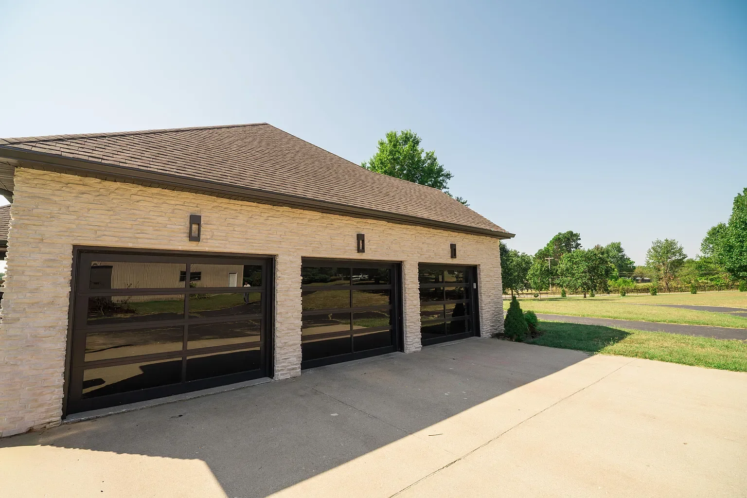 A modern garage with three black glass-paneled doors, beige brick exterior, and brown shingle roof in a grassy area with trees in the background.