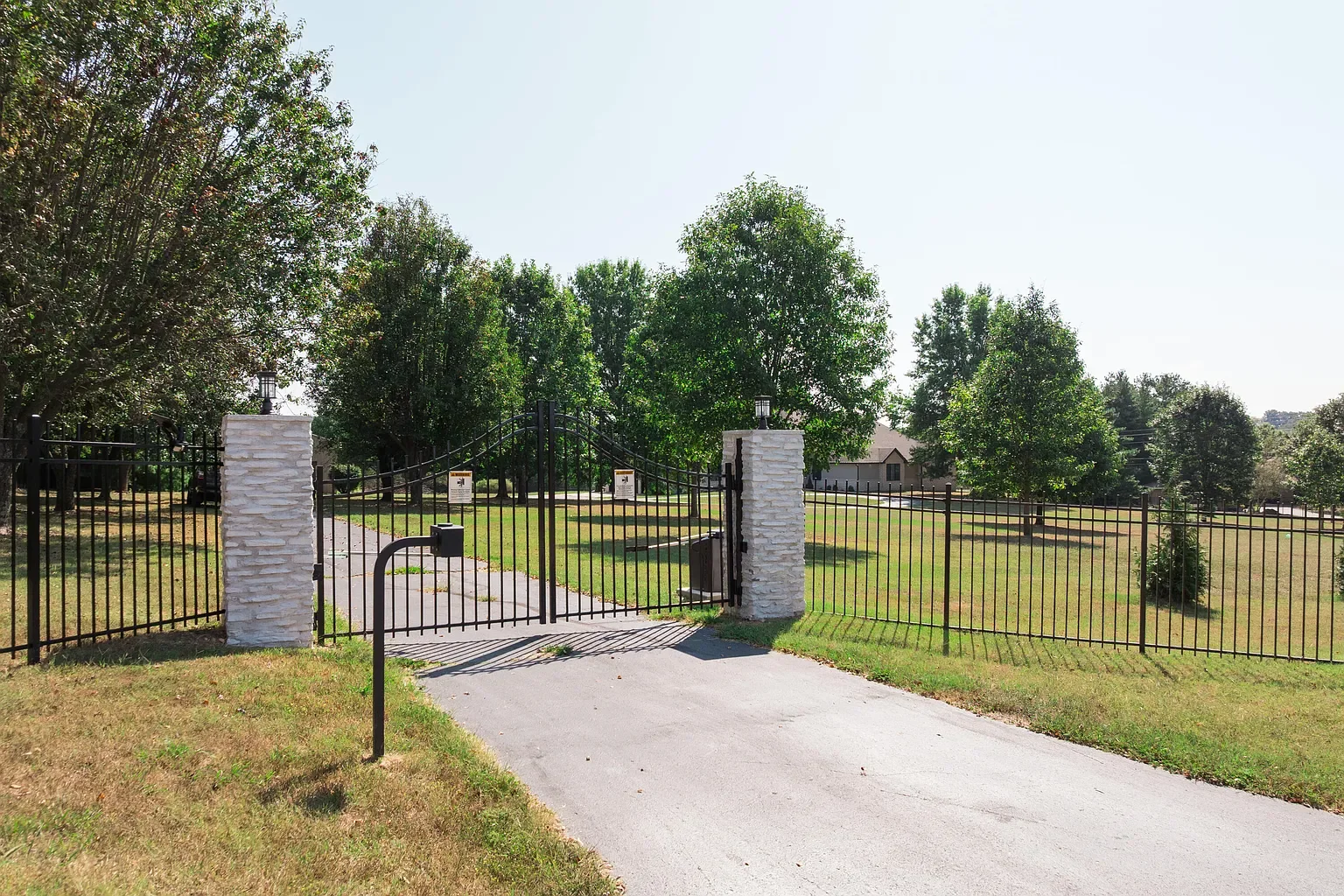 A black metal gate with two brick pillars on each side, leading into a grassy yard with trees in the background.