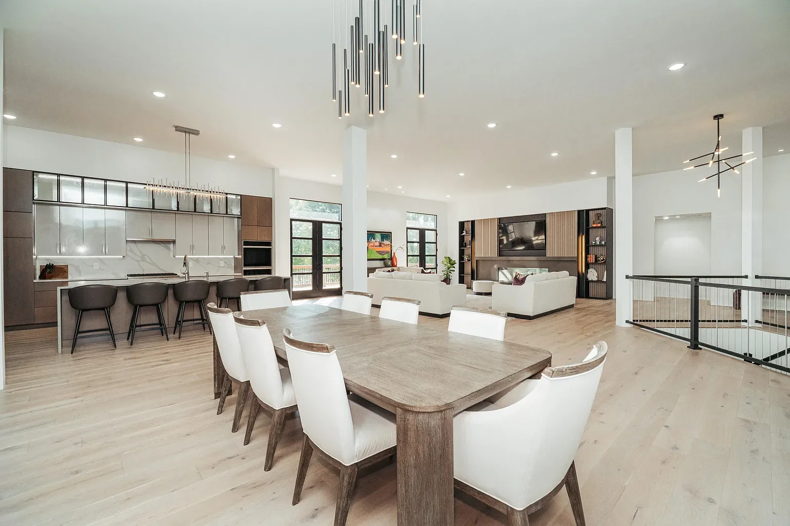 Open-concept living room and kitchen with modern decor. Includes dining table with chairs, kitchen island with bar stools, white sofas, built-in shelves, and large windows letting in natural light.