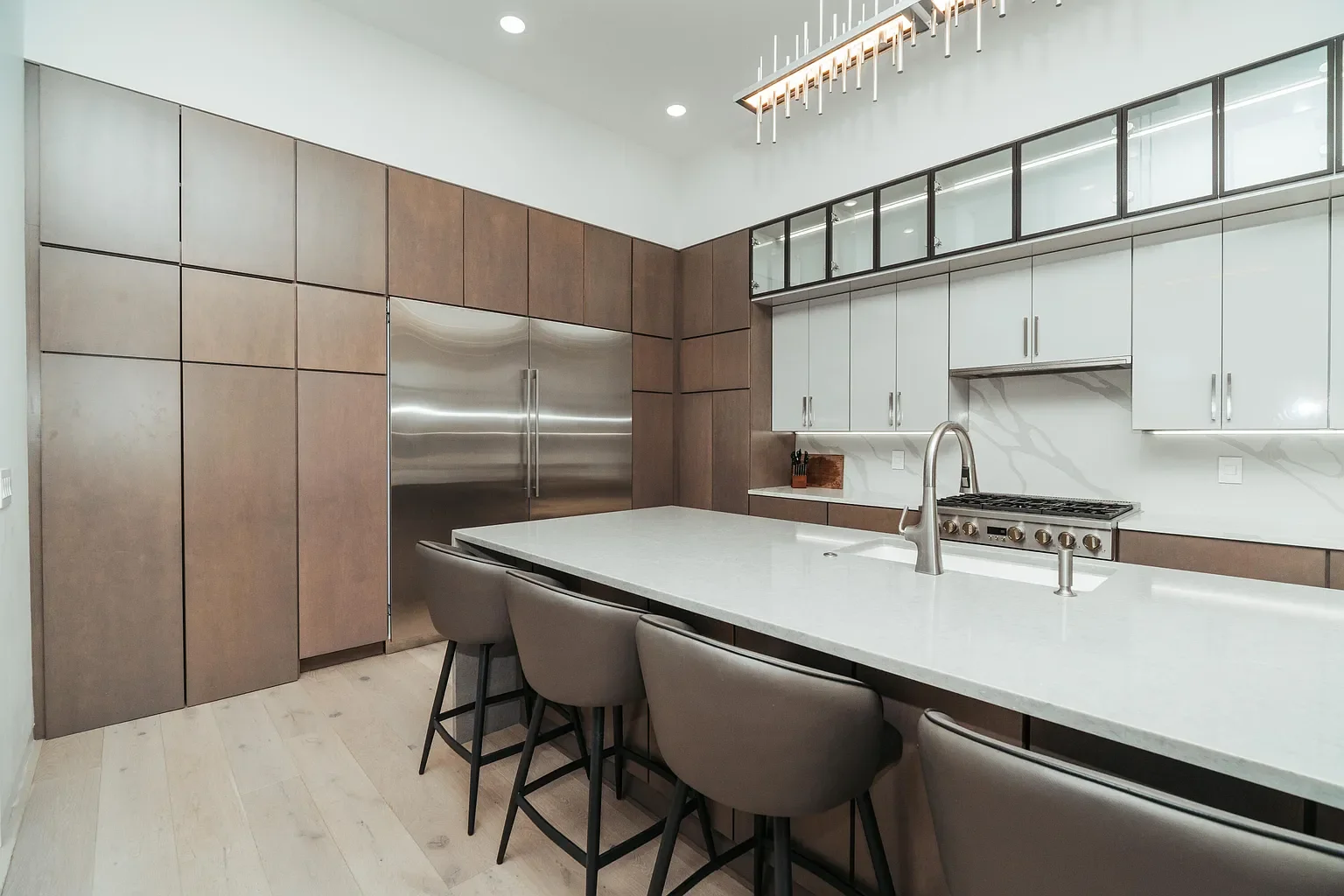 Modern kitchen with white countertops, gray bar stools, light wood flooring, stainless steel appliances, and upper glass-front cabinets.