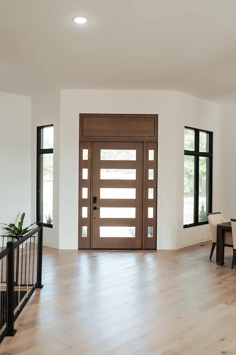 Modern front door with horizontal glass panels, flanked by two tall windows, in a bright entryway with light wood flooring and minimal decor.