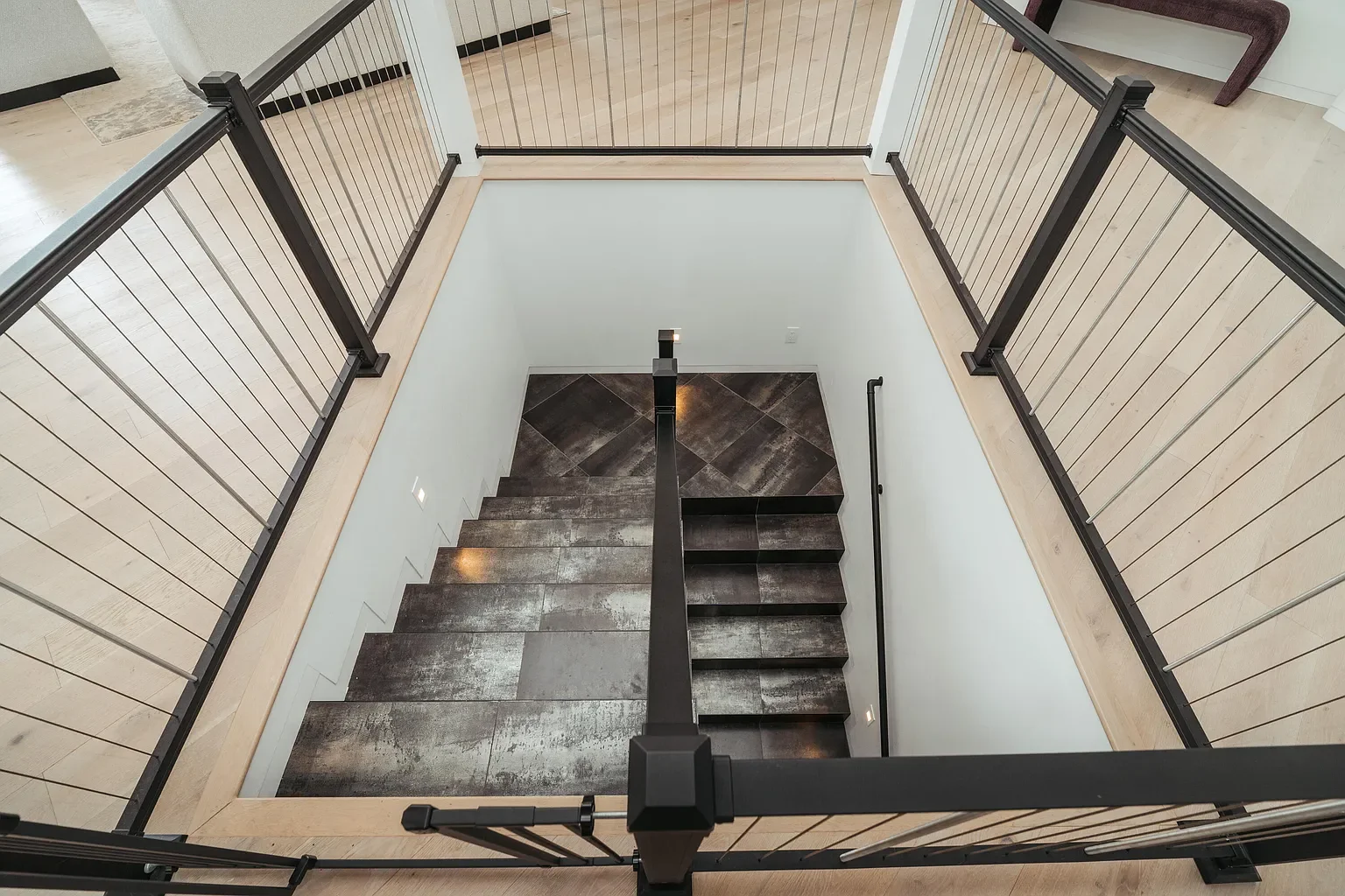 View looking down a staircase with black handrails and cables, dark tiled steps, and a landing area with black and gray tiles, surrounded by white walls and a wooden floor.
