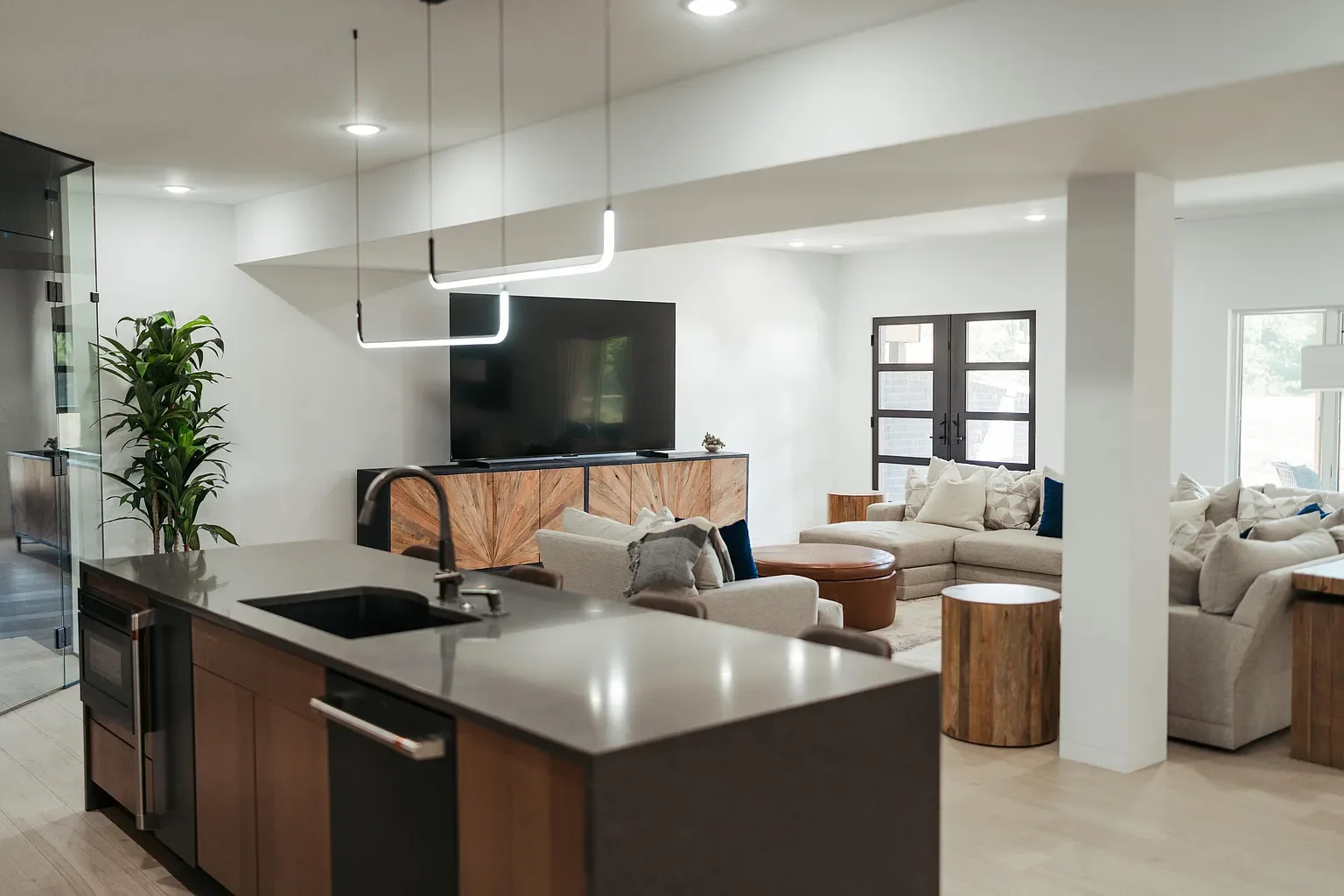 Open-concept living room and kitchen with beige sofas, wooden side tables, large flat-screen TV on a wooden console, potted plant, and modern pendant lights, with a sliding glass door and windows allowing natural light.