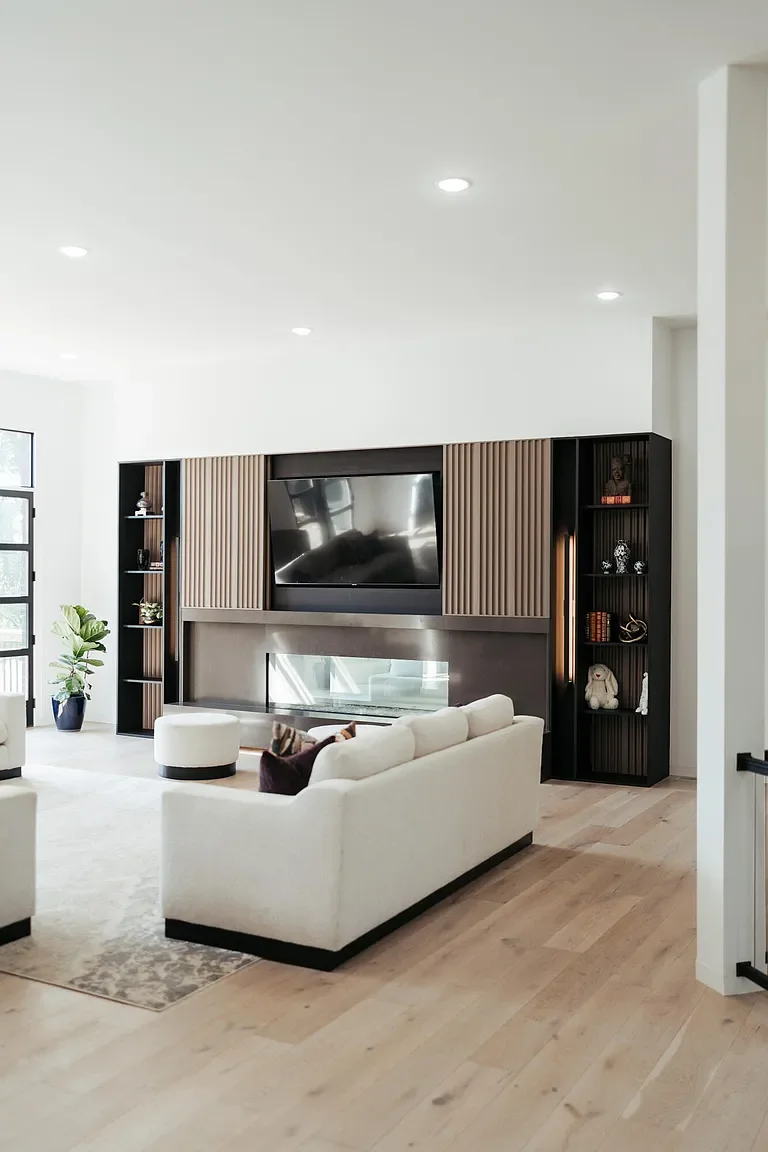 Modern living room with white sectional sofa, black and wood shelving, large TV mounted above a sleek fireplace, light hardwood floors, and a potted plant near the window.