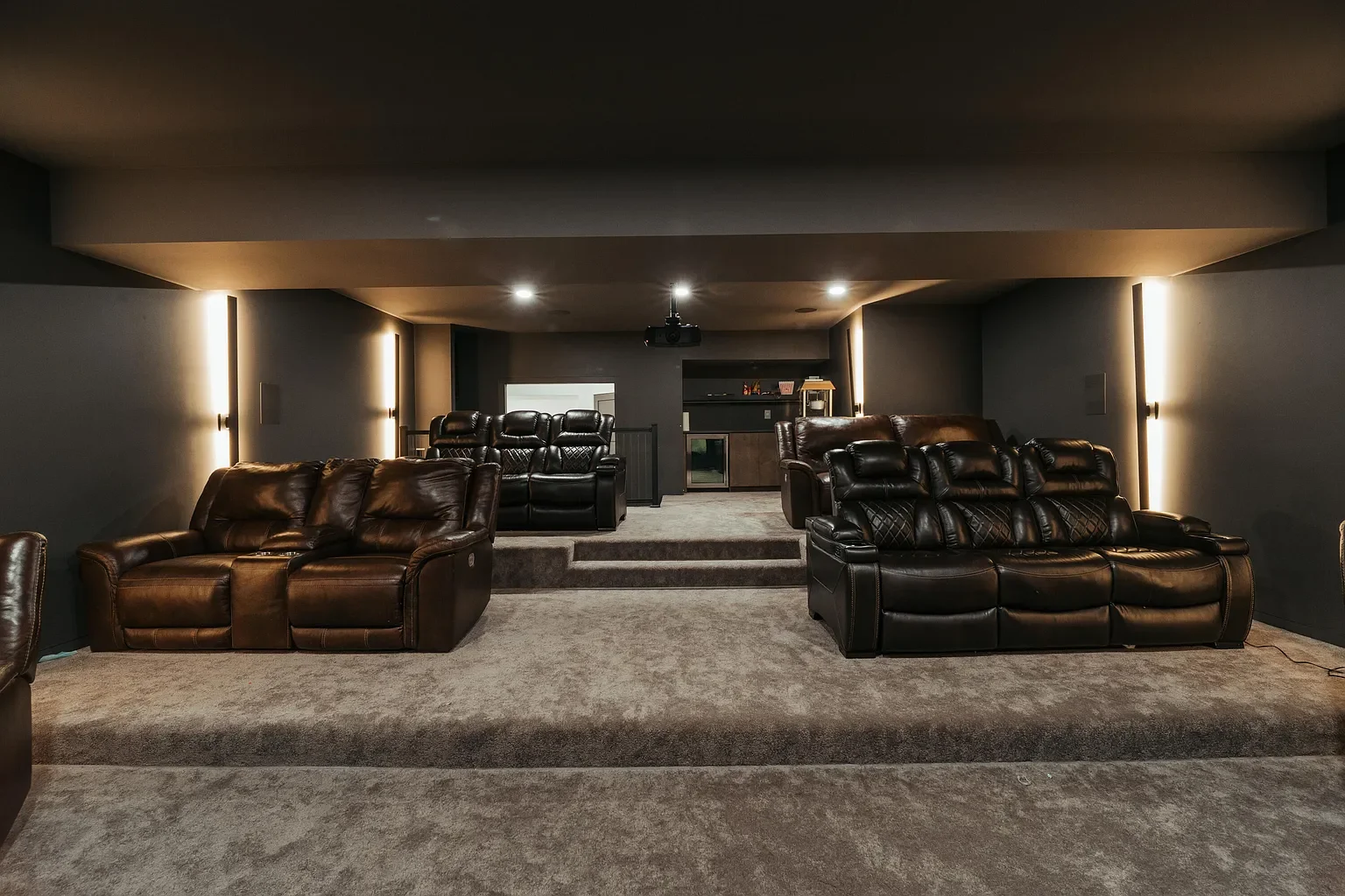 Basement home theater with a large gray carpeted floor, three brown leather sofas, and two black leather recliners, with a projector mounted on the ceiling, dim lighting, and a screen or window at the back.