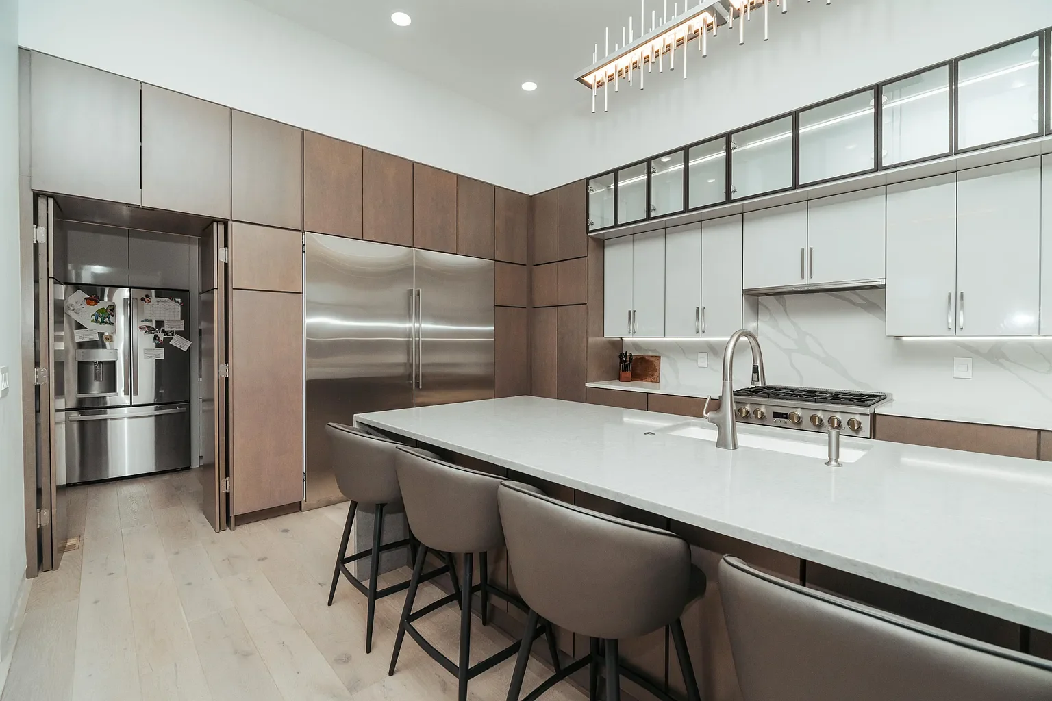 Modern kitchen with a large white island, gray bar stools, stainless steel refrigerator, built-in stove, and upper cabinets with glass door sections.
