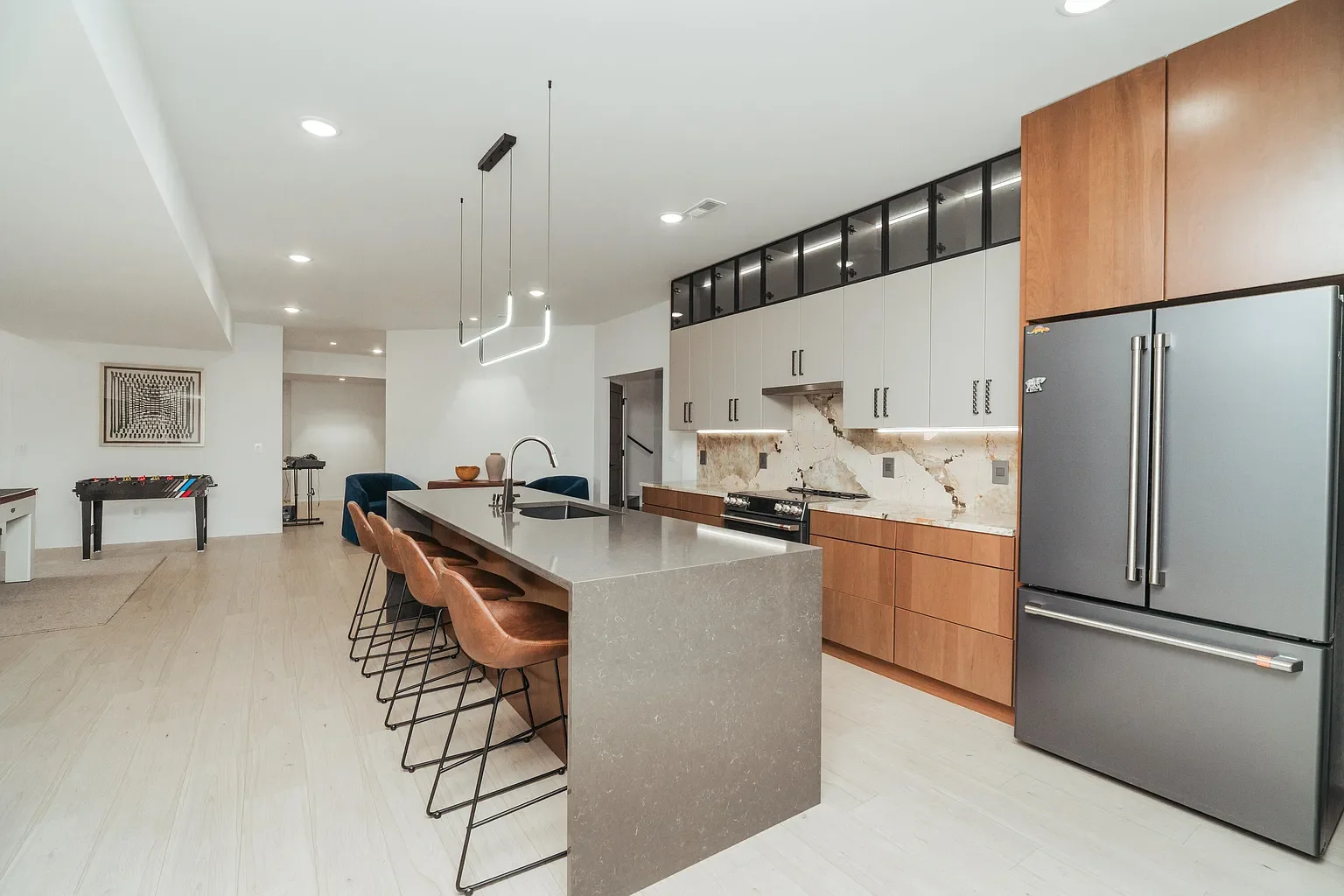 Modern kitchen with a large island, bar stools, wood and white cabinets, stainless steel refrigerator, marble backsplash, and contemporary lighting.