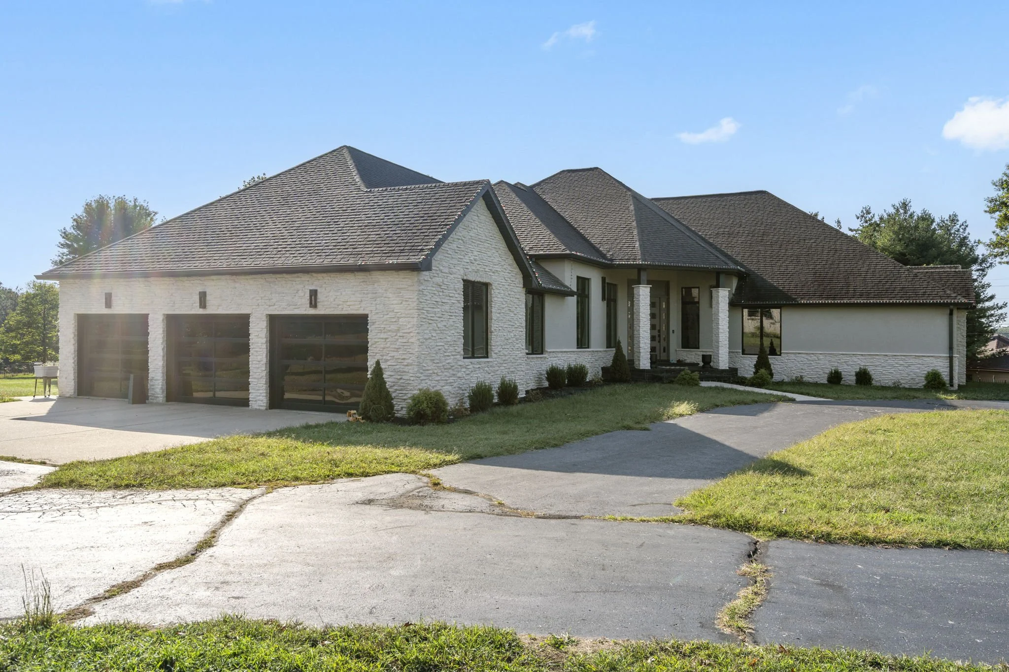 Front view of a modern house with white brick exterior, dark shingles, and an attached three-car garage. The driveway is cracked and runs in front of the house, with patches of grass on either side. There are small shrubs and trees along the house's foundation, under a clear blue sky.