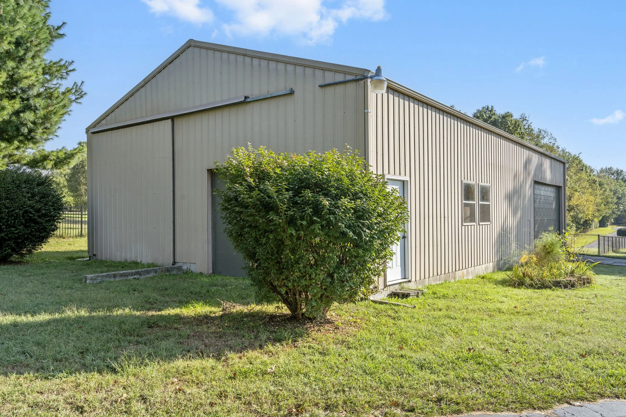 A beige metal building with a gable roof, a white door, two small windows, and a garage door, surrounded by green grass, bushes, and trees under a partly cloudy sky.
