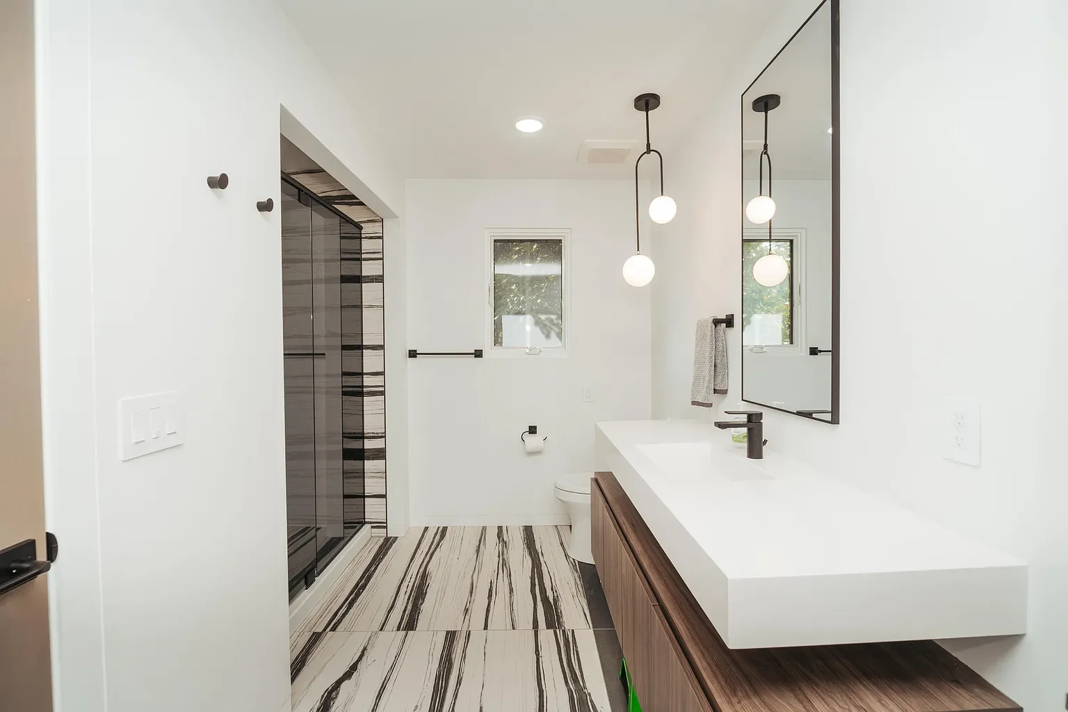 Modern and minimalistic bathroom with white walls, a large mirror, black-framed pendant lights, wood vanity, marble floor, and a walk-in shower with striped tiles.