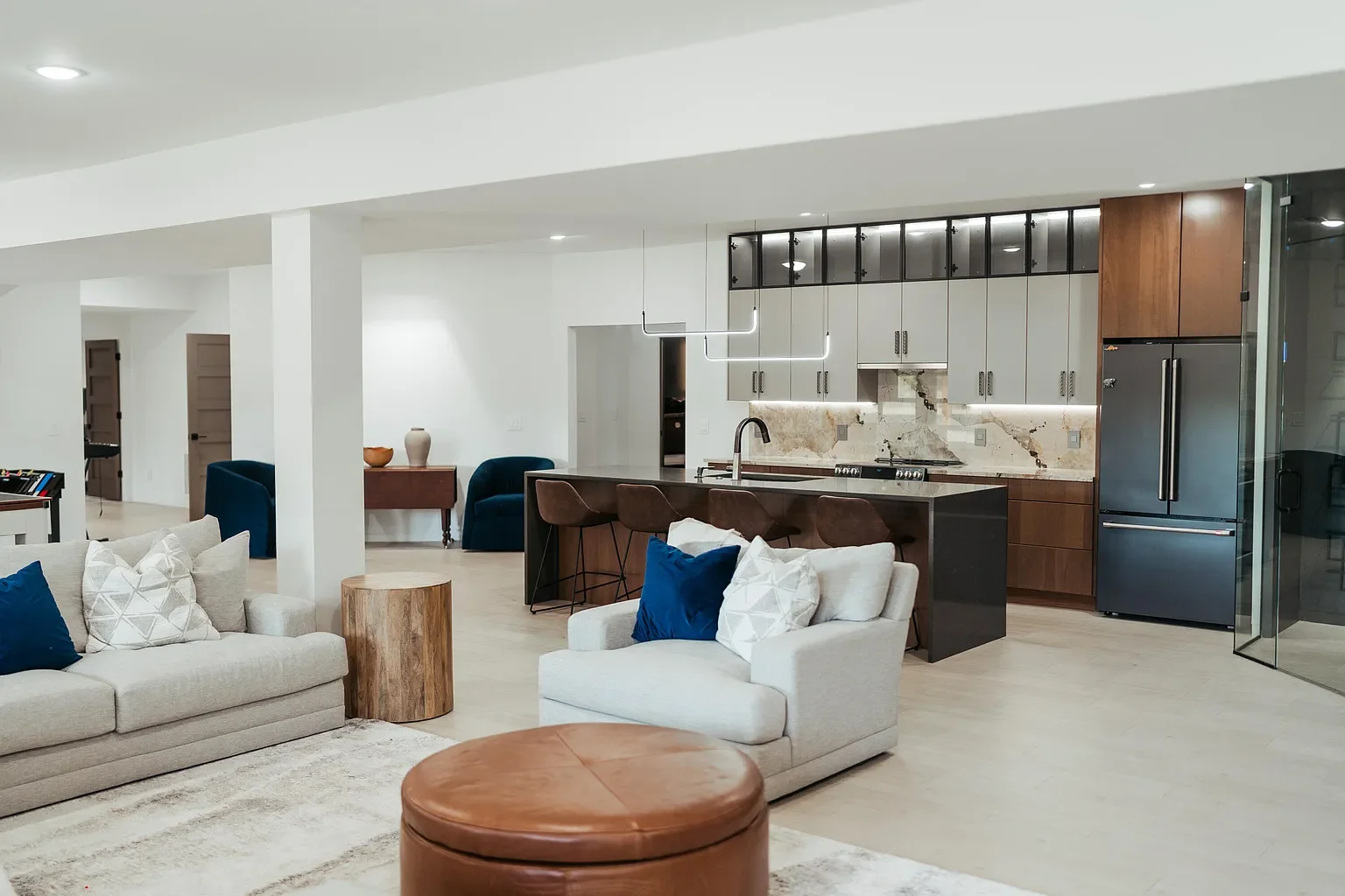 Open-concept living room and kitchen with modern decor, beige sofa, armchair with pillows, wooden and leather accents, and a sleek kitchen with marble backsplash and stainless steel appliances.