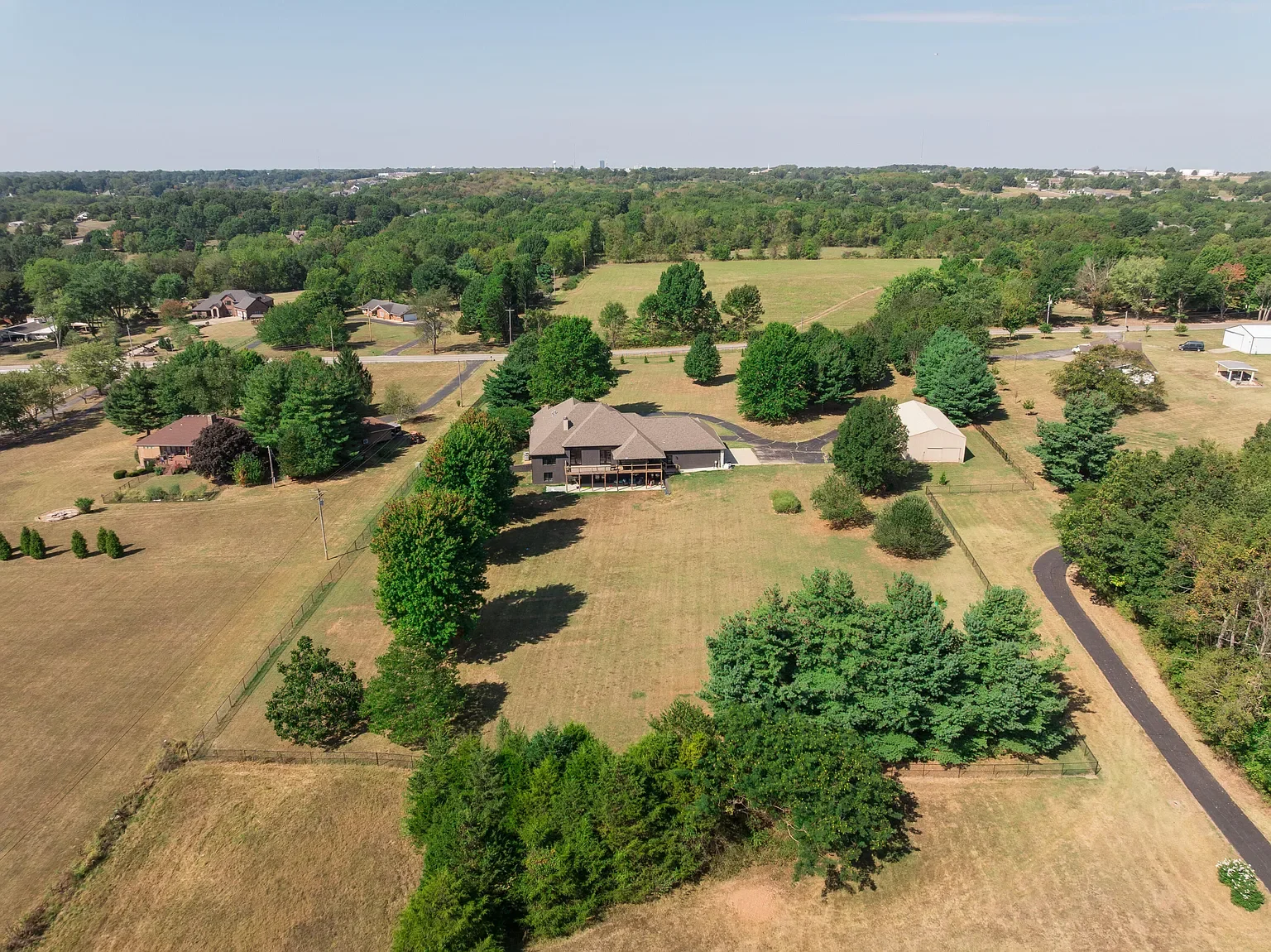 Aerial view of a large property with a house, trees, open grassy areas, and surrounding neighborhood in the distance.
