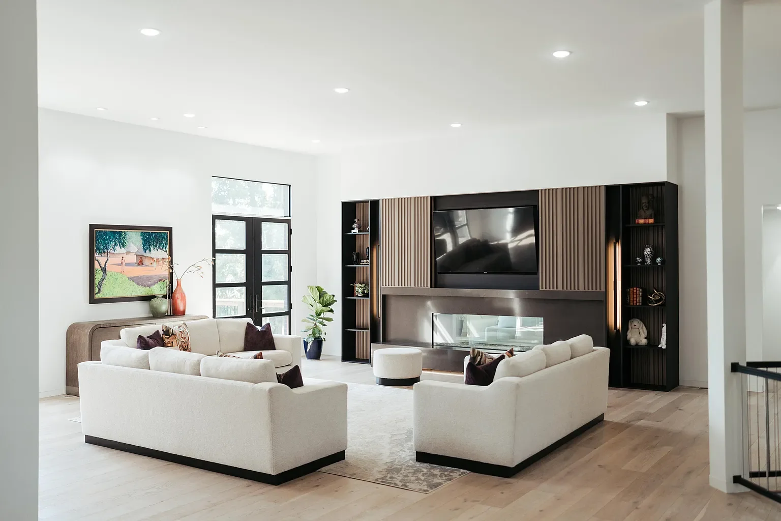 Modern living room with white sofas, a large wall-mounted television, a fireplace, and decorative shelves, with large windows and light wood flooring.