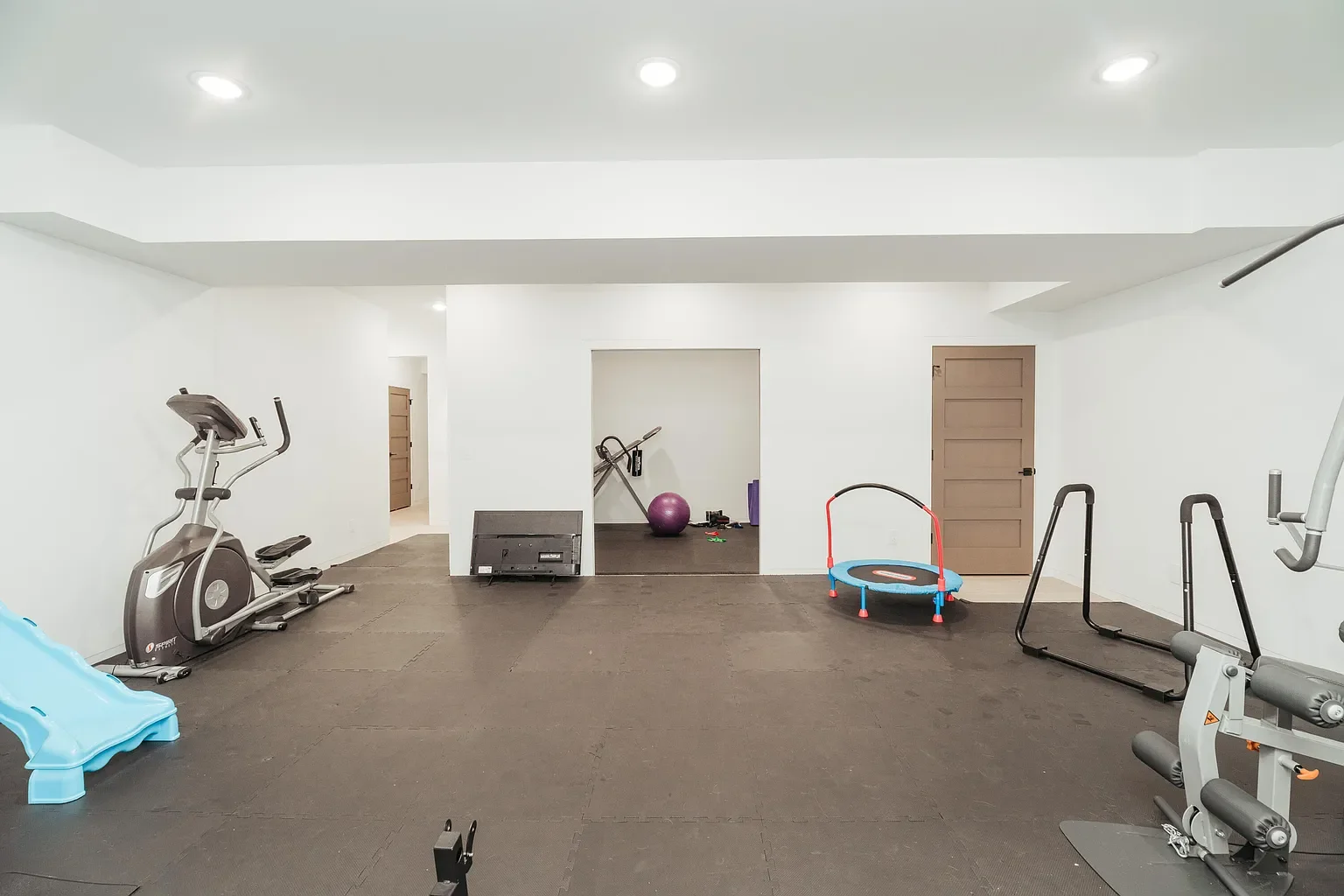 Empty home gym with exercise equipment including a treadmill, elliptical, small trampoline, stability ball, American football, and foam mats on the floor.