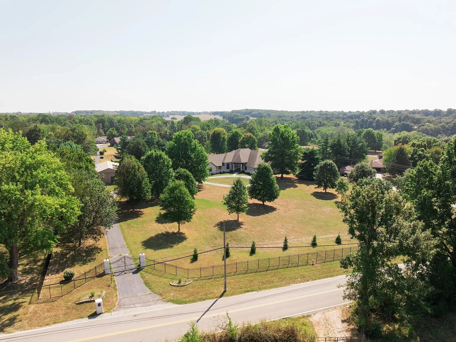 A large house with a gray roof and white walls is surrounded by numerous green trees in a spacious yard. A curved driveway leads to the house, with a smaller walkway connecting to a gated entrance on the main road. A wooden fence encloses part of the yard, and there is a utility pole with power lines nearby. The background consists of an expansive green landscape with more trees and a clear sky.