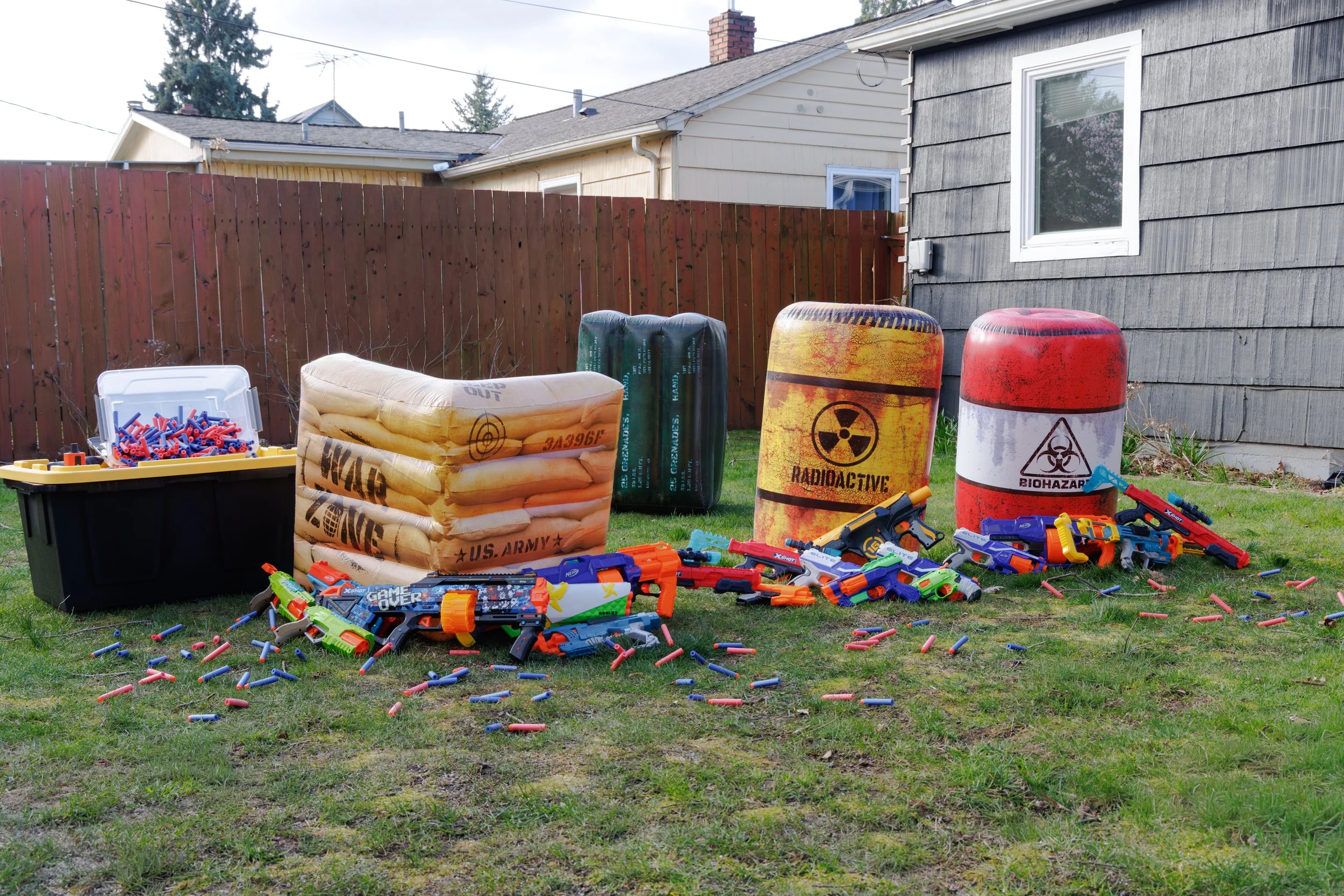 Backyard scene with toy foam dart guns, scattered foam darts, and five large barrels labeled with hazardous materials symbols, including radioactive and biohazard, set against a house and a wooden fence.