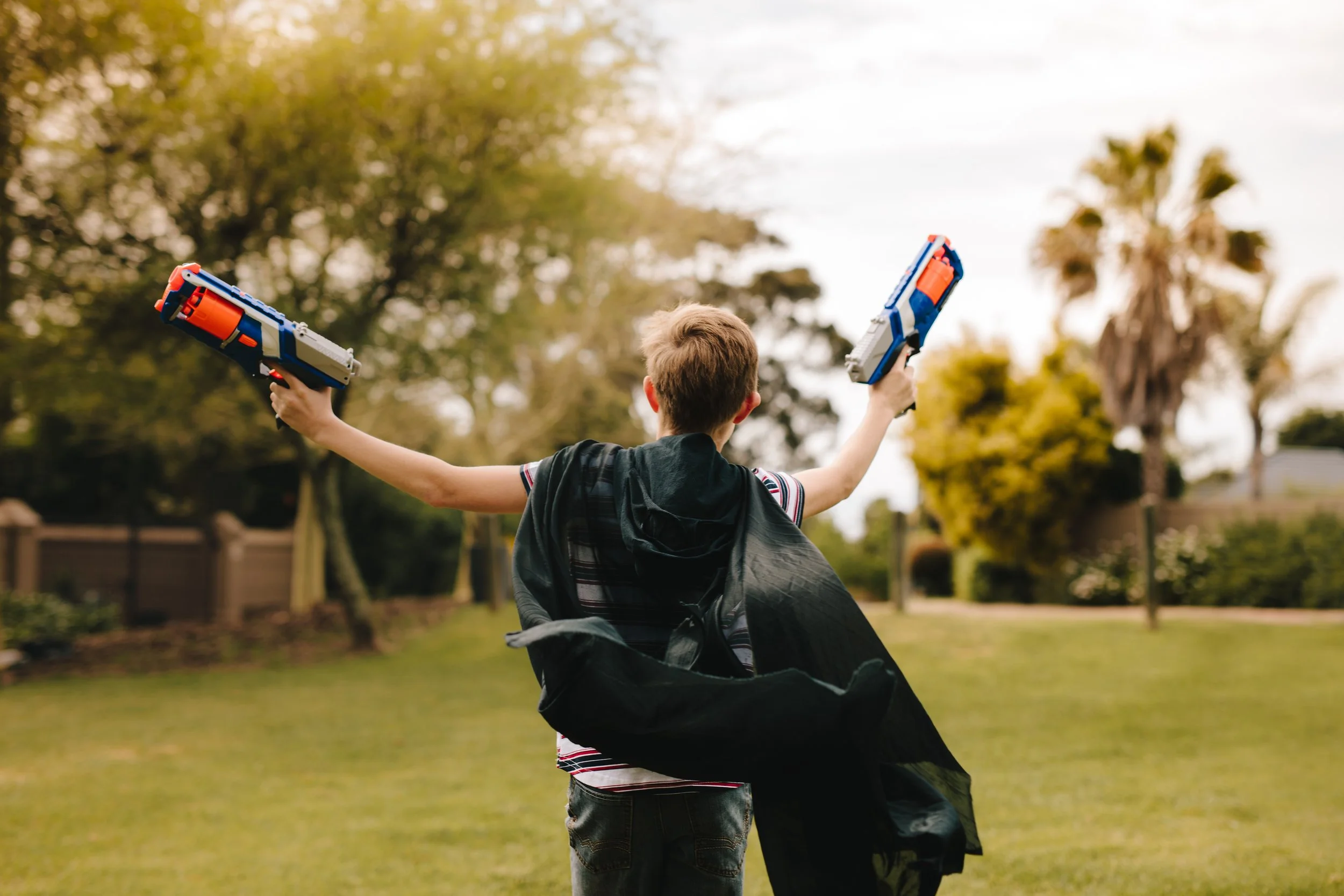 A boy holding two toy water guns with arms outstretched outdoors in a grassy backyard, with trees and a wooden fence in the background.