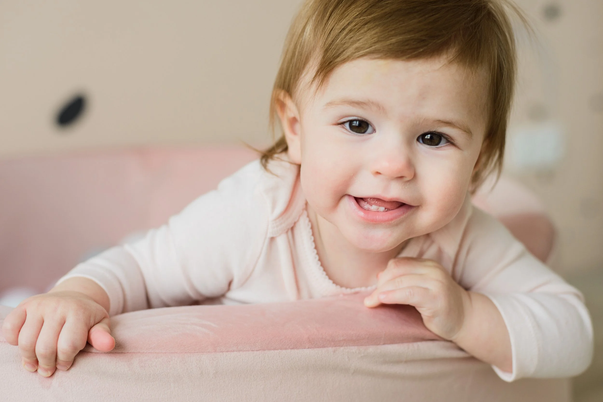Close-up of a smiling toddler with light brown hair, lying on a pink cushioned surface, wearing a light-colored long sleeve shirt.