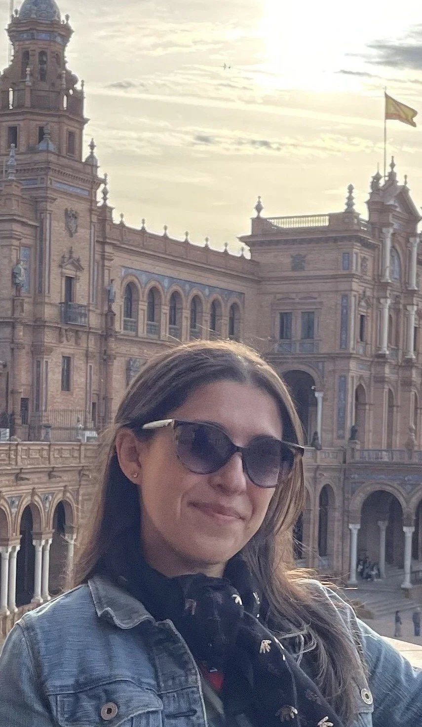 A woman wearing sunglasses and a denim jacket standing in front of an ornate historic building.