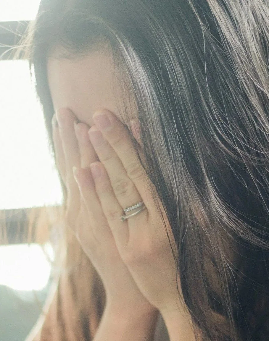 A woman with long dark hair covering her face with her hands, showing a wedding ring on her left ring finger.