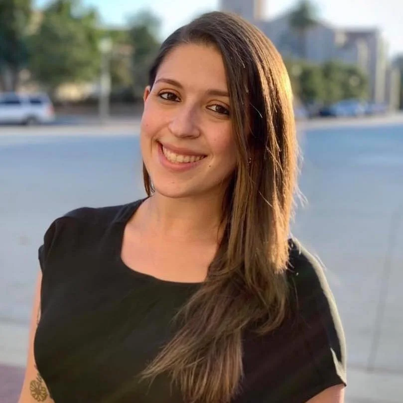 A young woman with long brown hair, smiling and wearing a black top, standing outdoors with blurred trees, cars, and buildings in the background.
