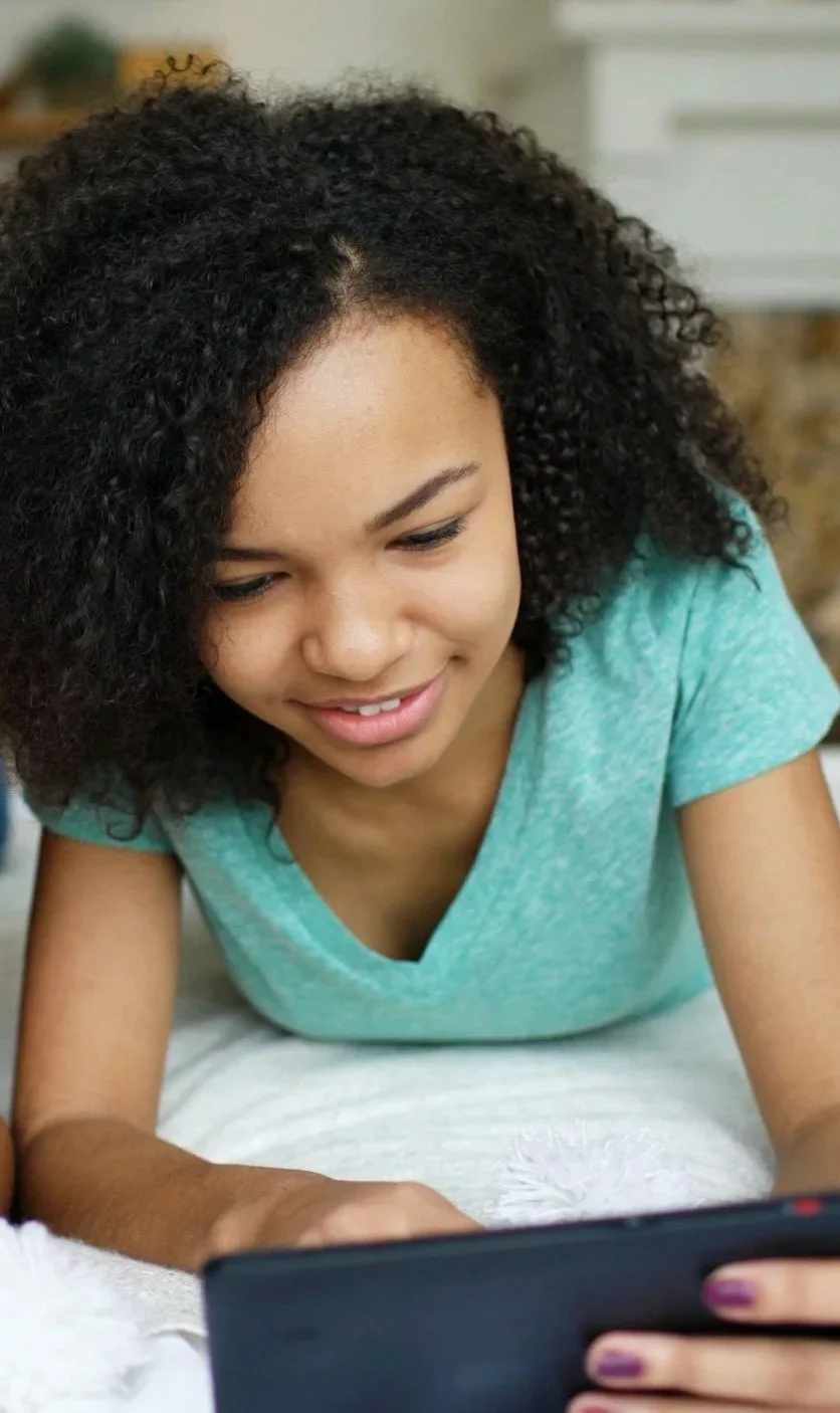 Young woman with curly hair lying on her stomach, using a tablet device, smiling.